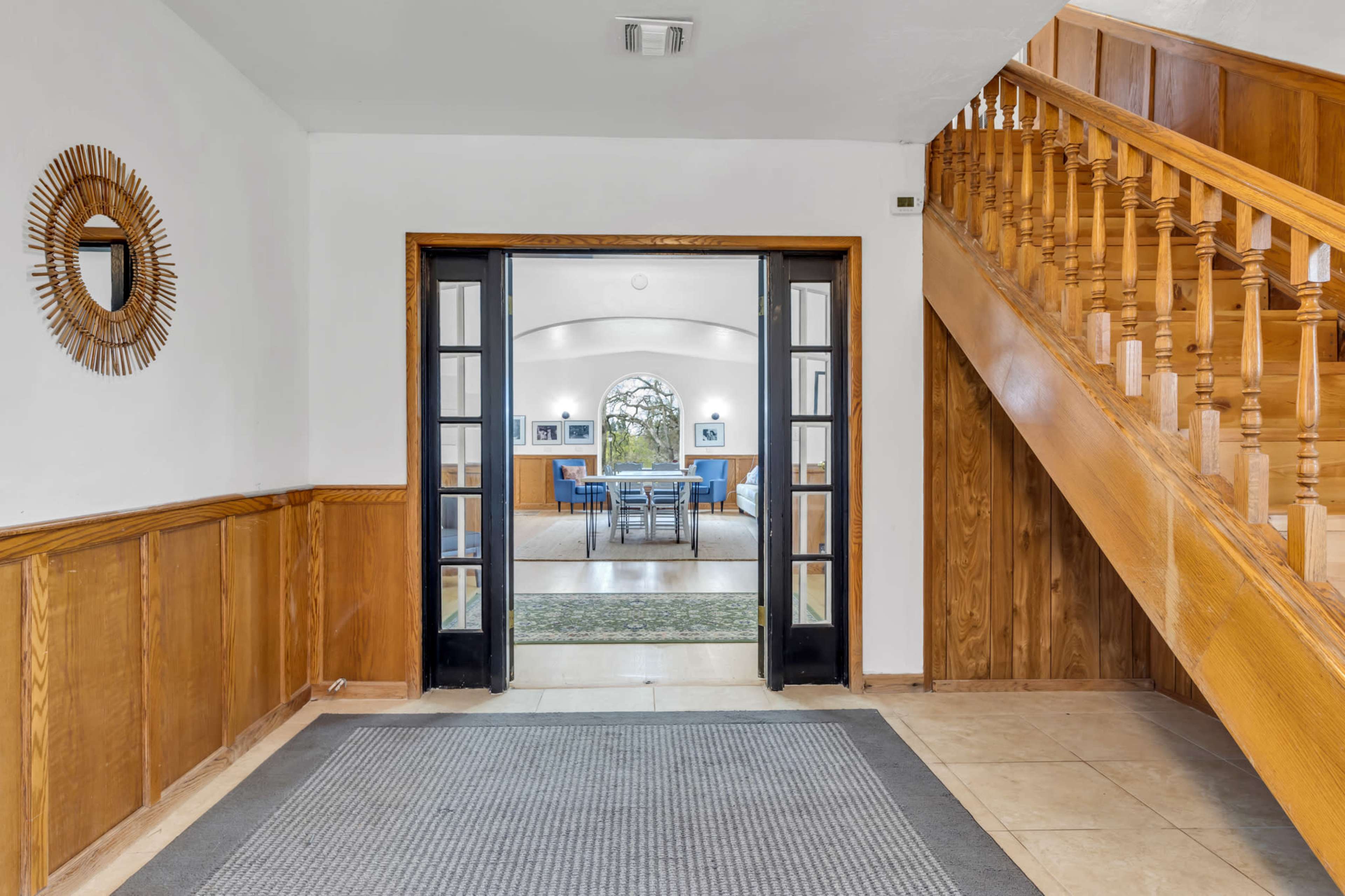 The image shows a foyer with a patterned rug, wooden walls, and a staircase leading to an upper level, framed by double doors that open into a dining area.