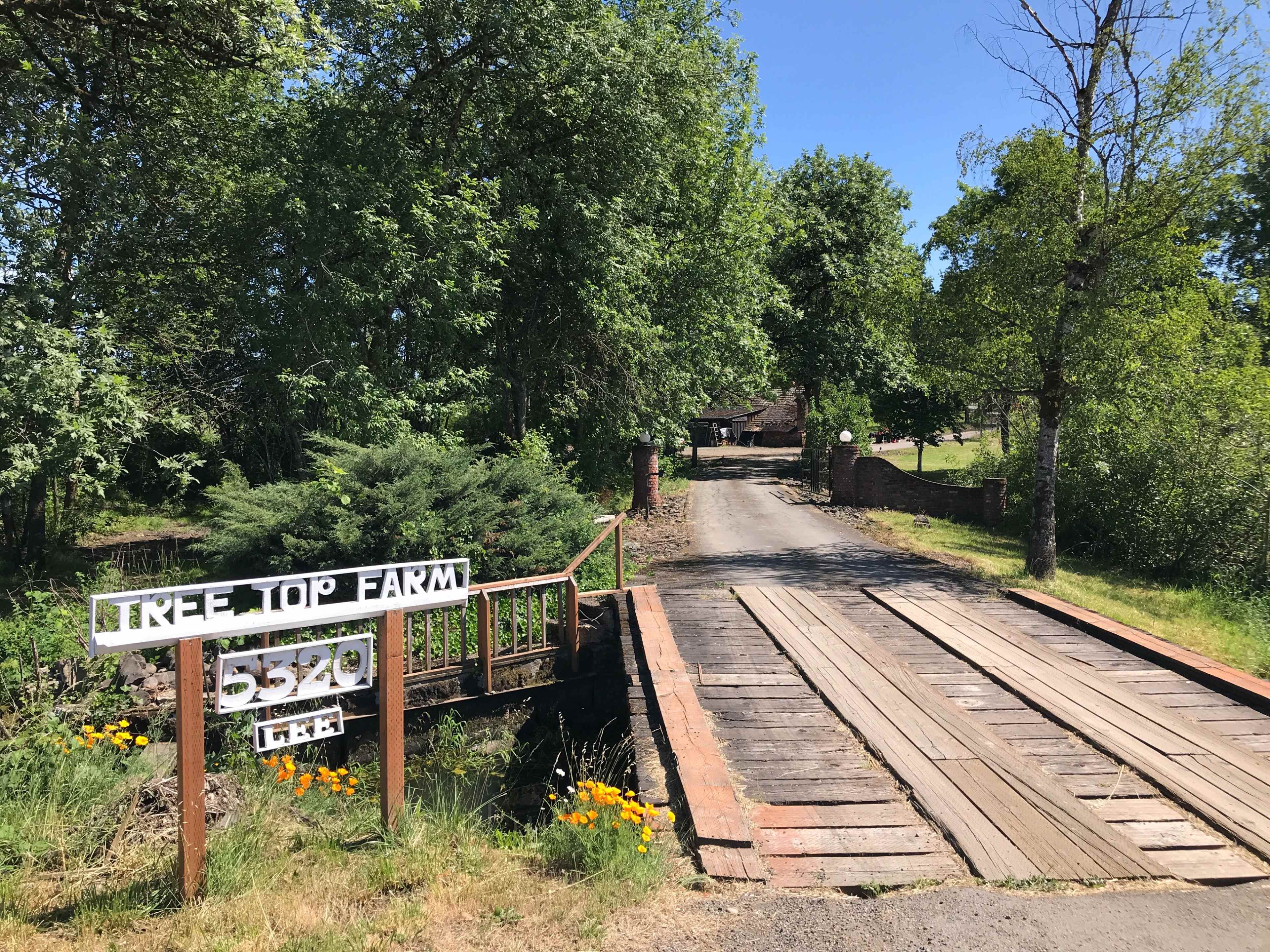 A wooden bridge leads to a dirt road flanked by greenery, with a sign indicating "Tree Top Farm" and the address 5220.