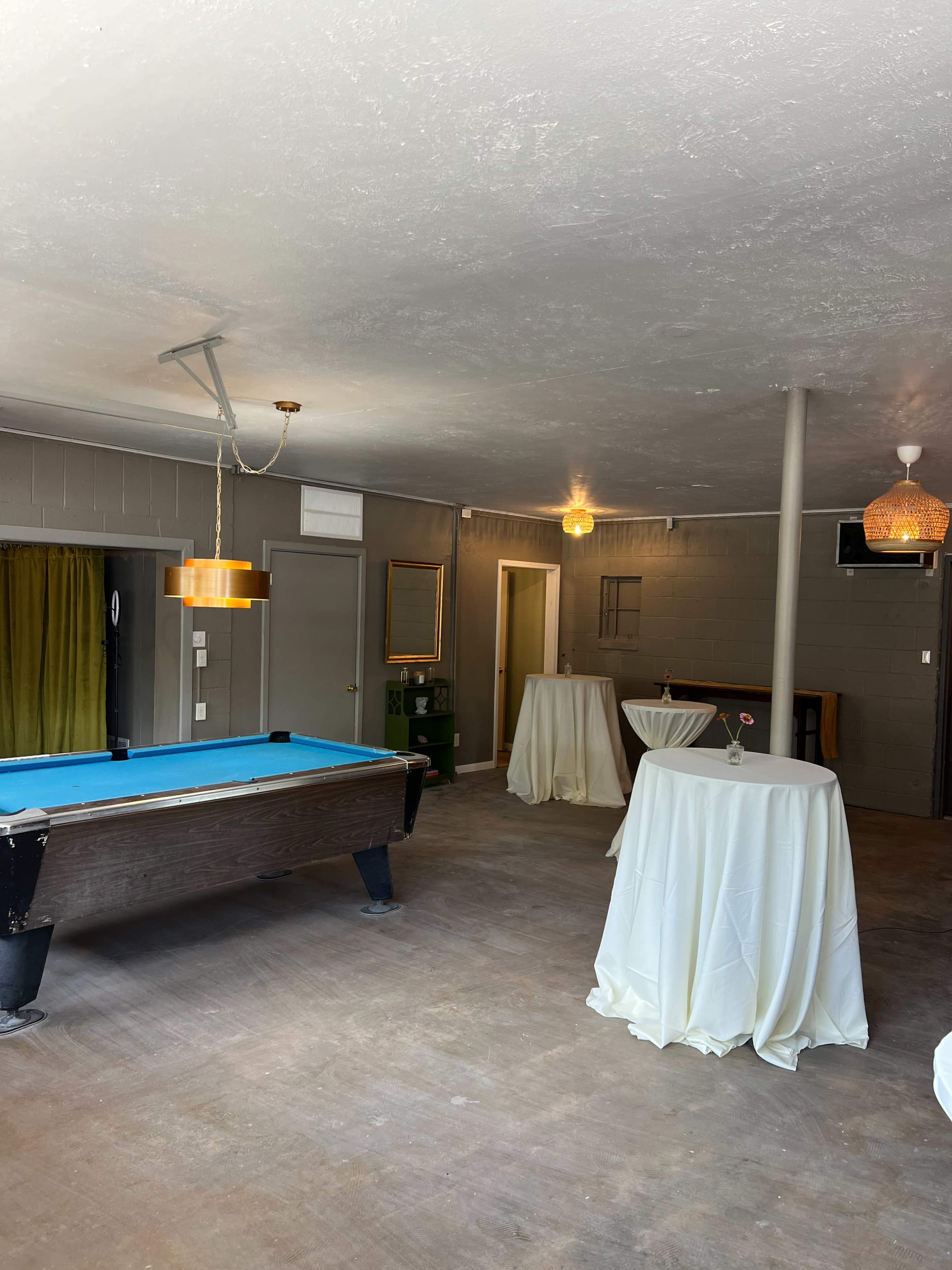 The image shows a spacious room featuring a billiards table, several draped tables, and a mix of overhead lighting fixtures.