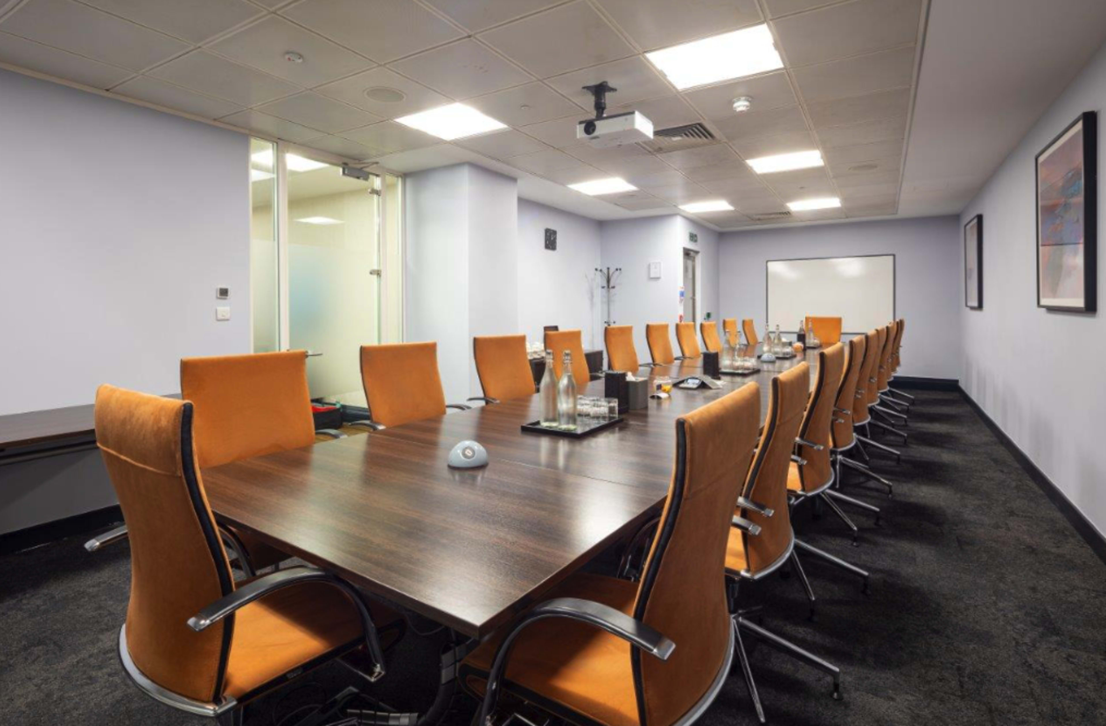A large conference room features a long wooden table surrounded by orange chairs, with a projector and whiteboard visible in the background.