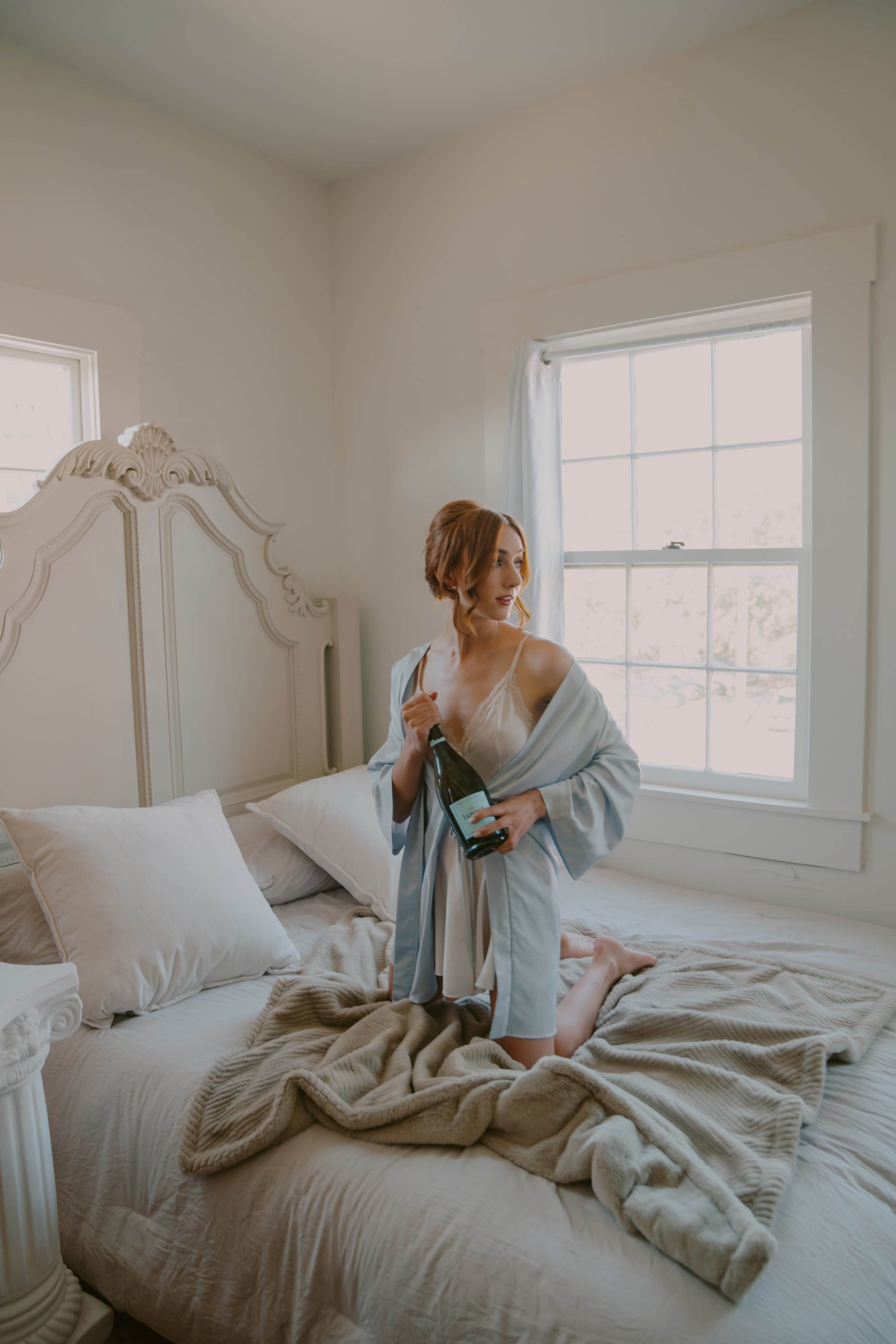 A woman stands on a bed in a well-lit room, holding a bottle of champagne while wrapped in a light-colored robe.