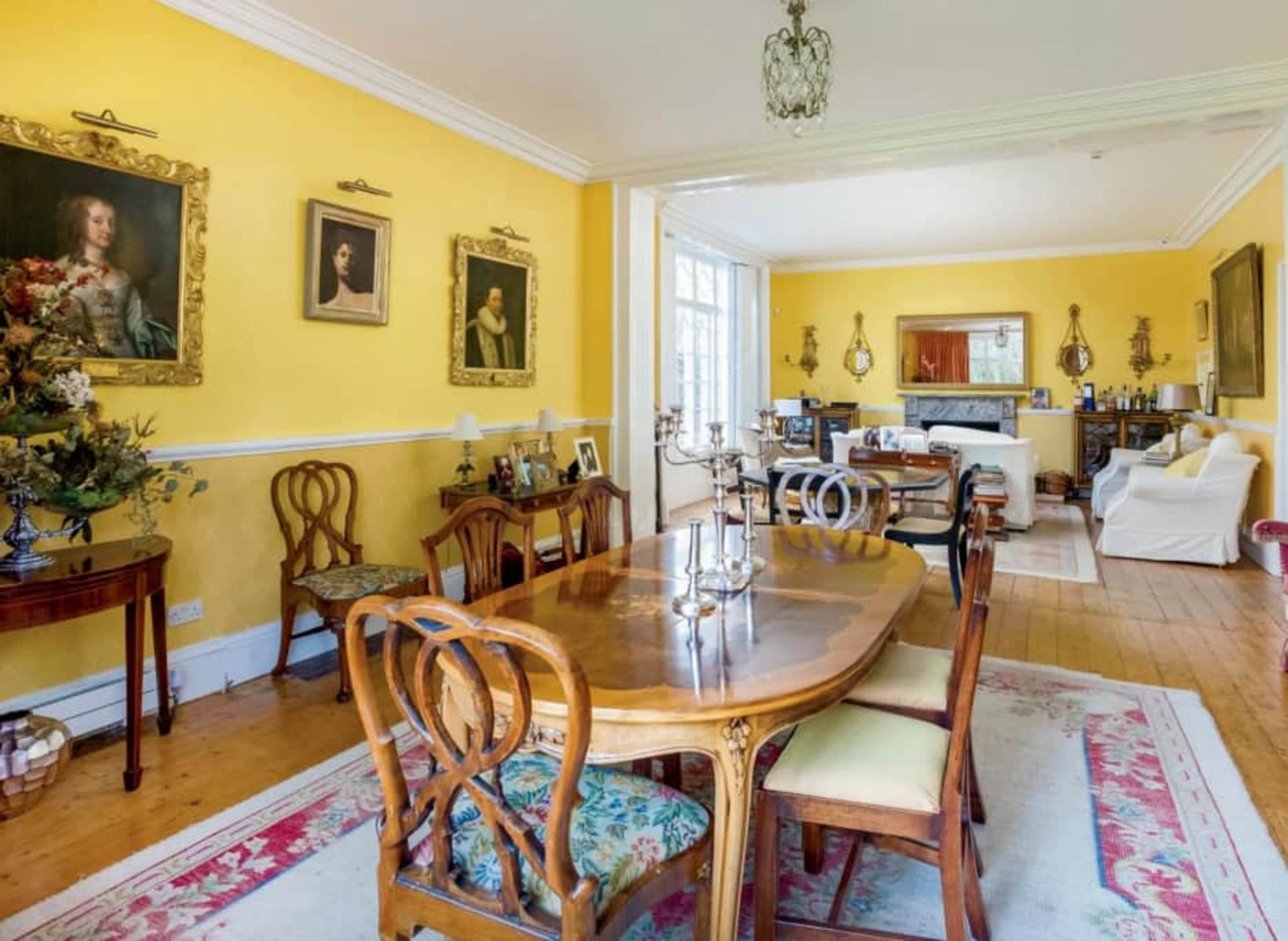 A bright dining room with yellow walls, a wooden table surrounded by ornate chairs, and framed portraits on the walls.