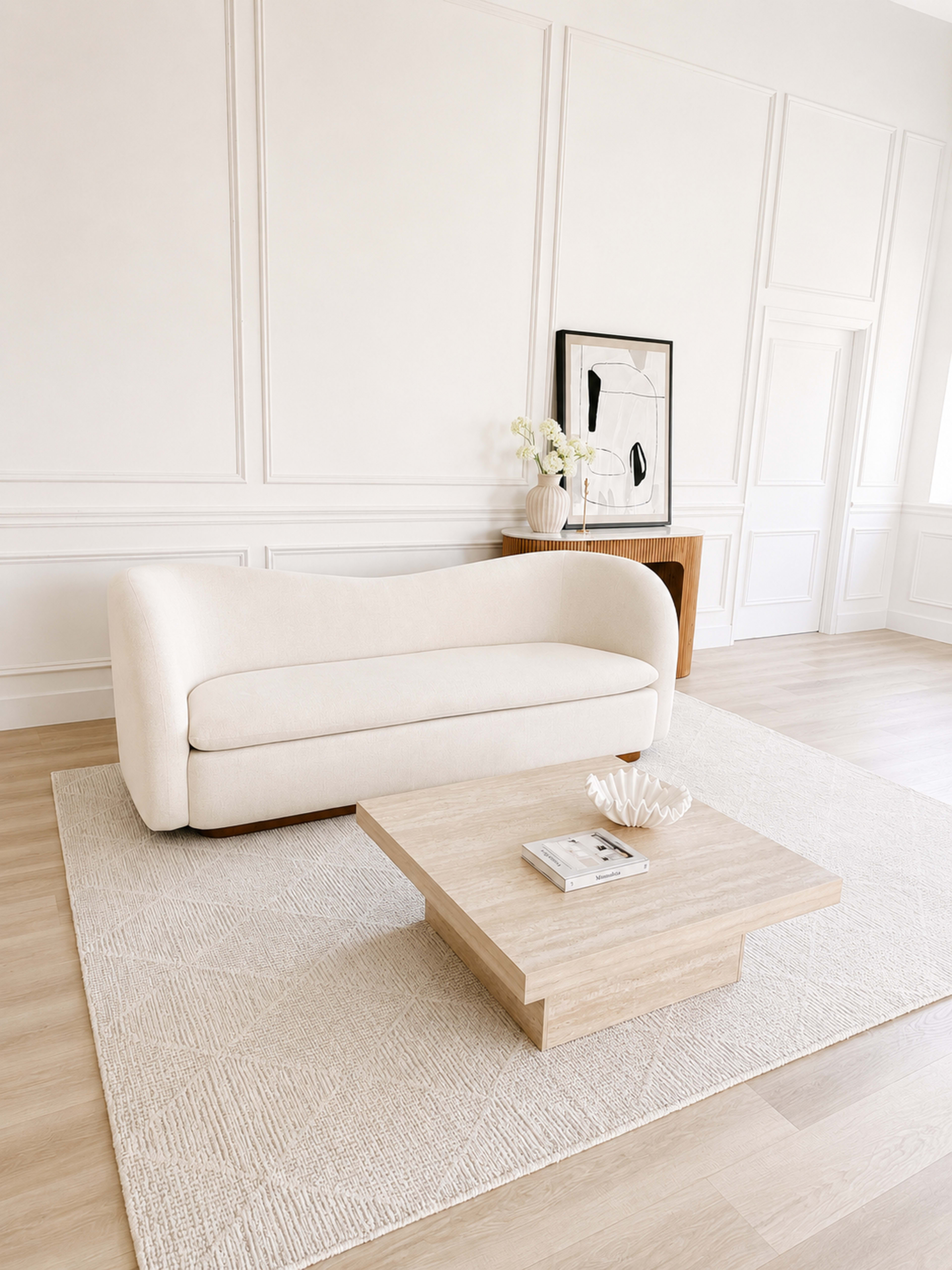 A spacious, minimalist living room features a large white sofa, a square wooden coffee table, and a decorative centerpiece on a light area rug.