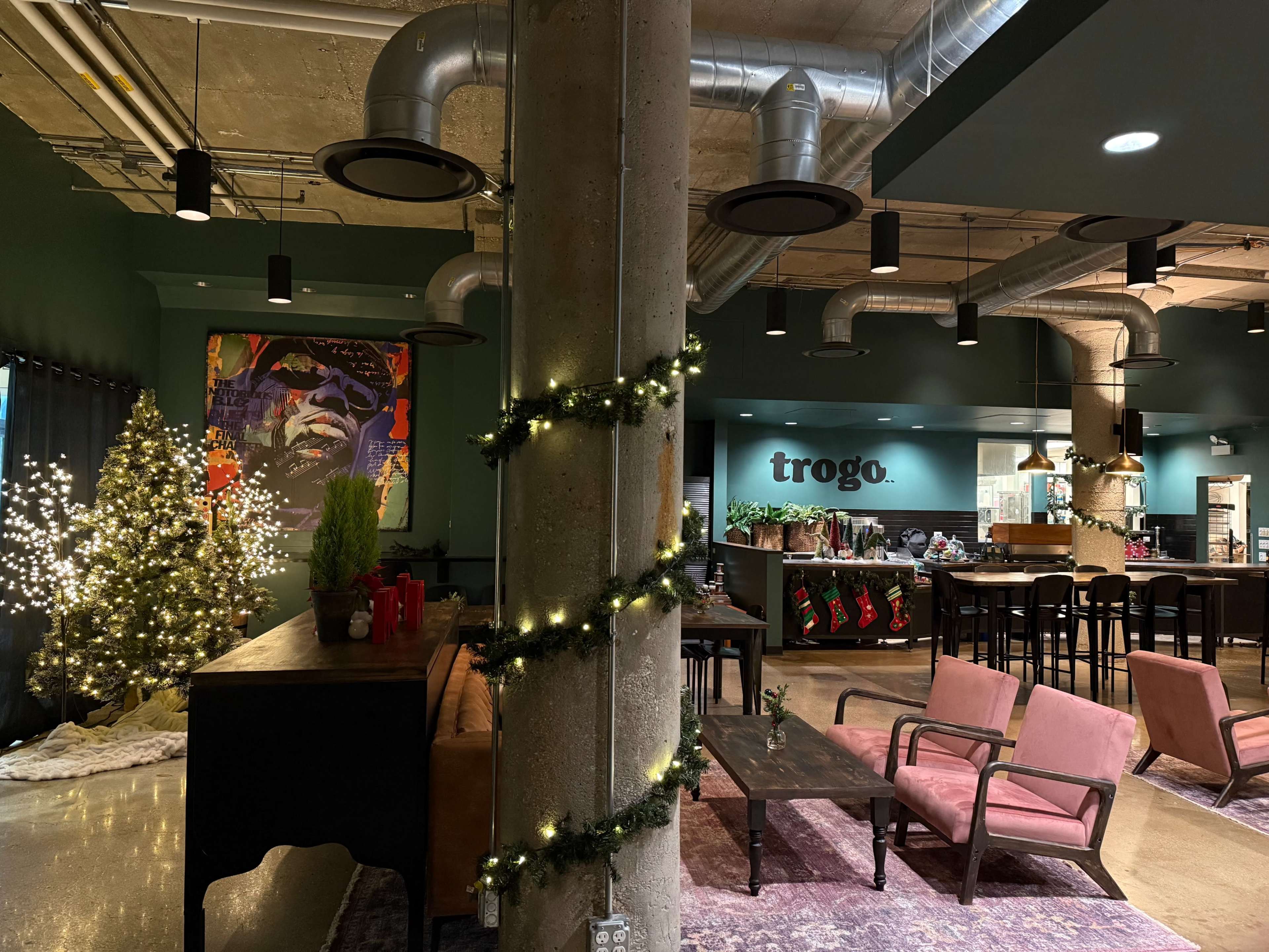 The image depicts a modern indoor space featuring a combination of seating areas, holiday decorations, and a bar, with a Christmas tree and artwork on the walls.
