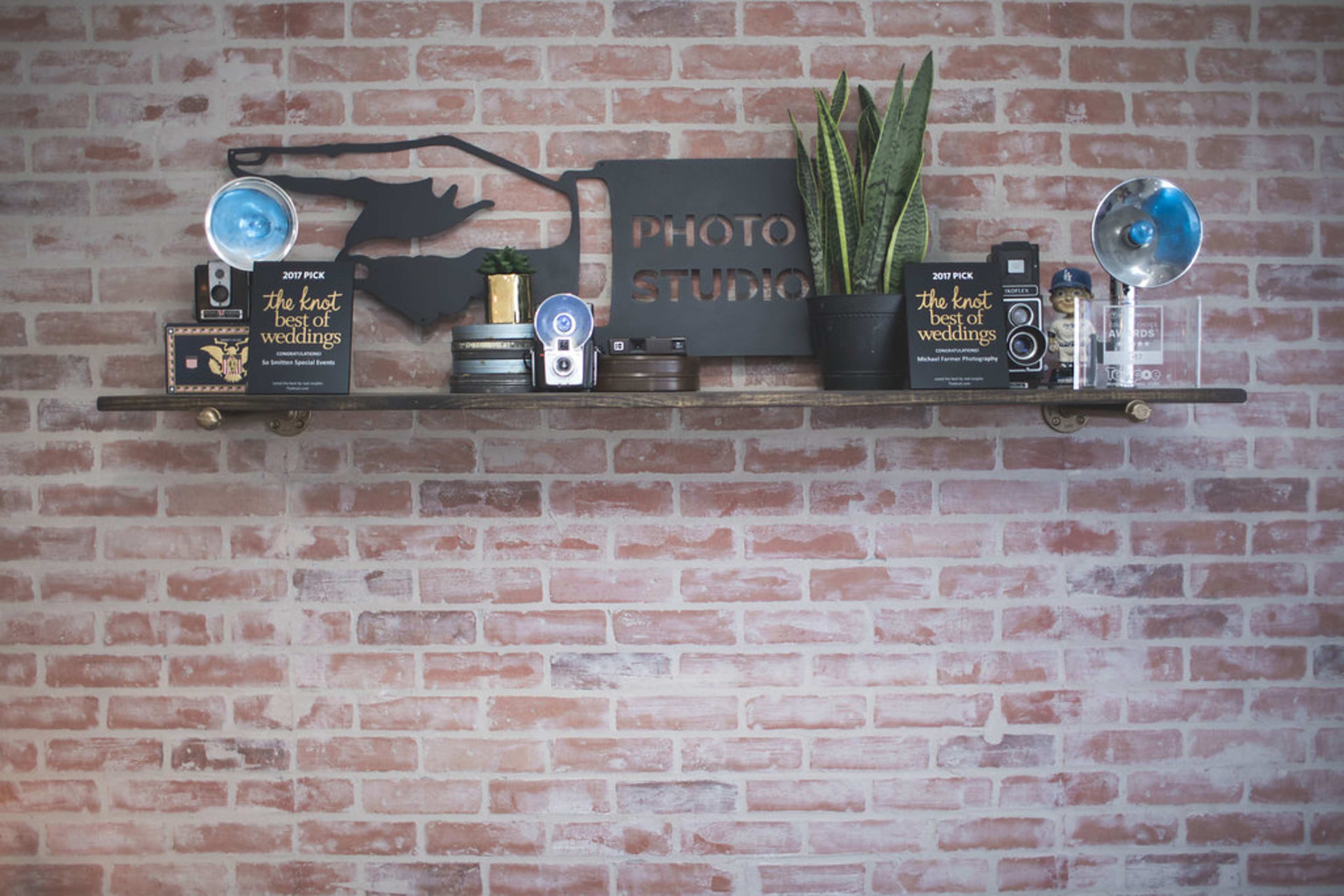 A wooden shelf displays various cameras, a plant, and decorative items against a brick wall with a "Photo Studio" sign.