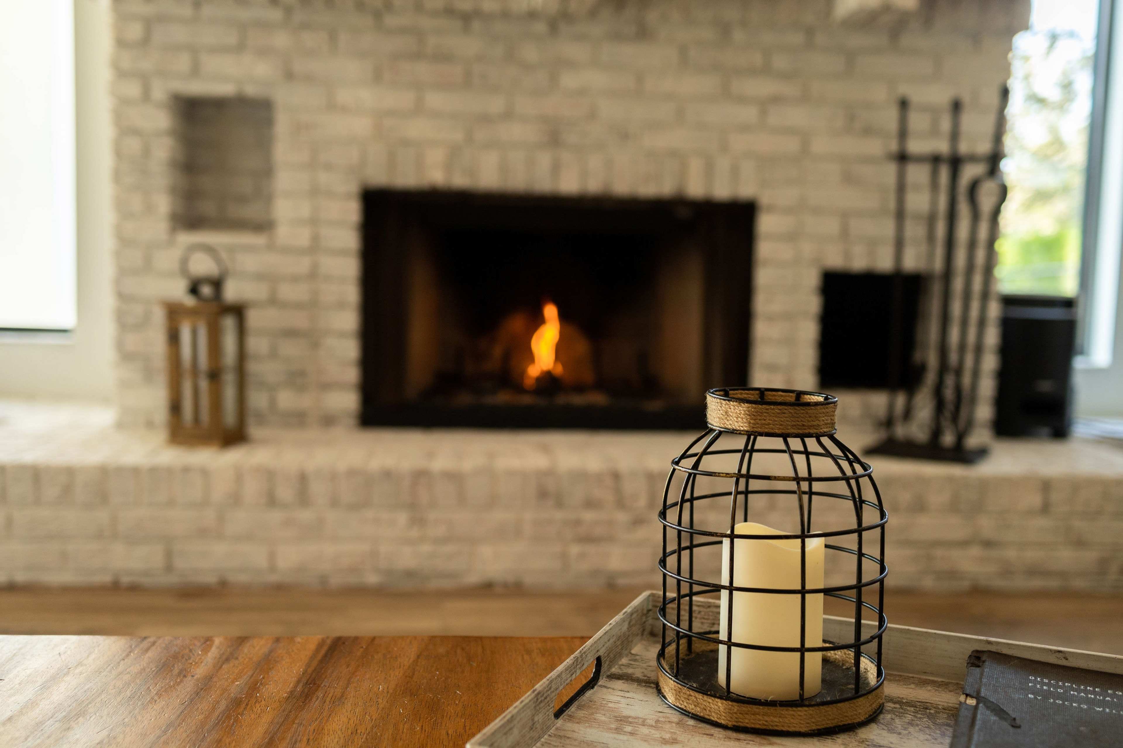 A candle in a metal cage sits on a tray in front of a brick fireplace with a small flame.