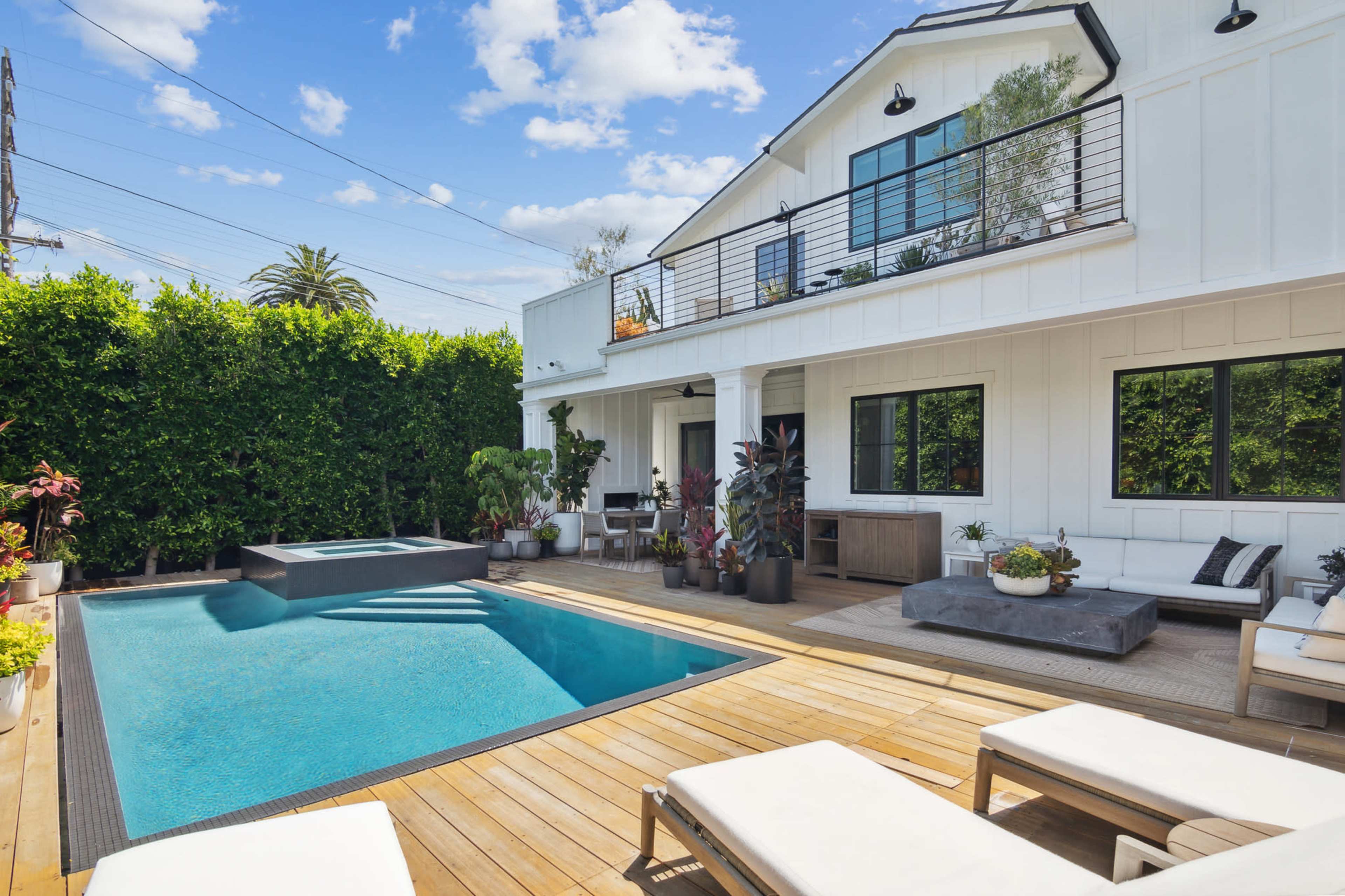 A modern outdoor area featuring a rectangular swimming pool surrounded by wooden decking, potted plants, and lounge chairs, alongside a white house with large windows and a second-floor balcony.
