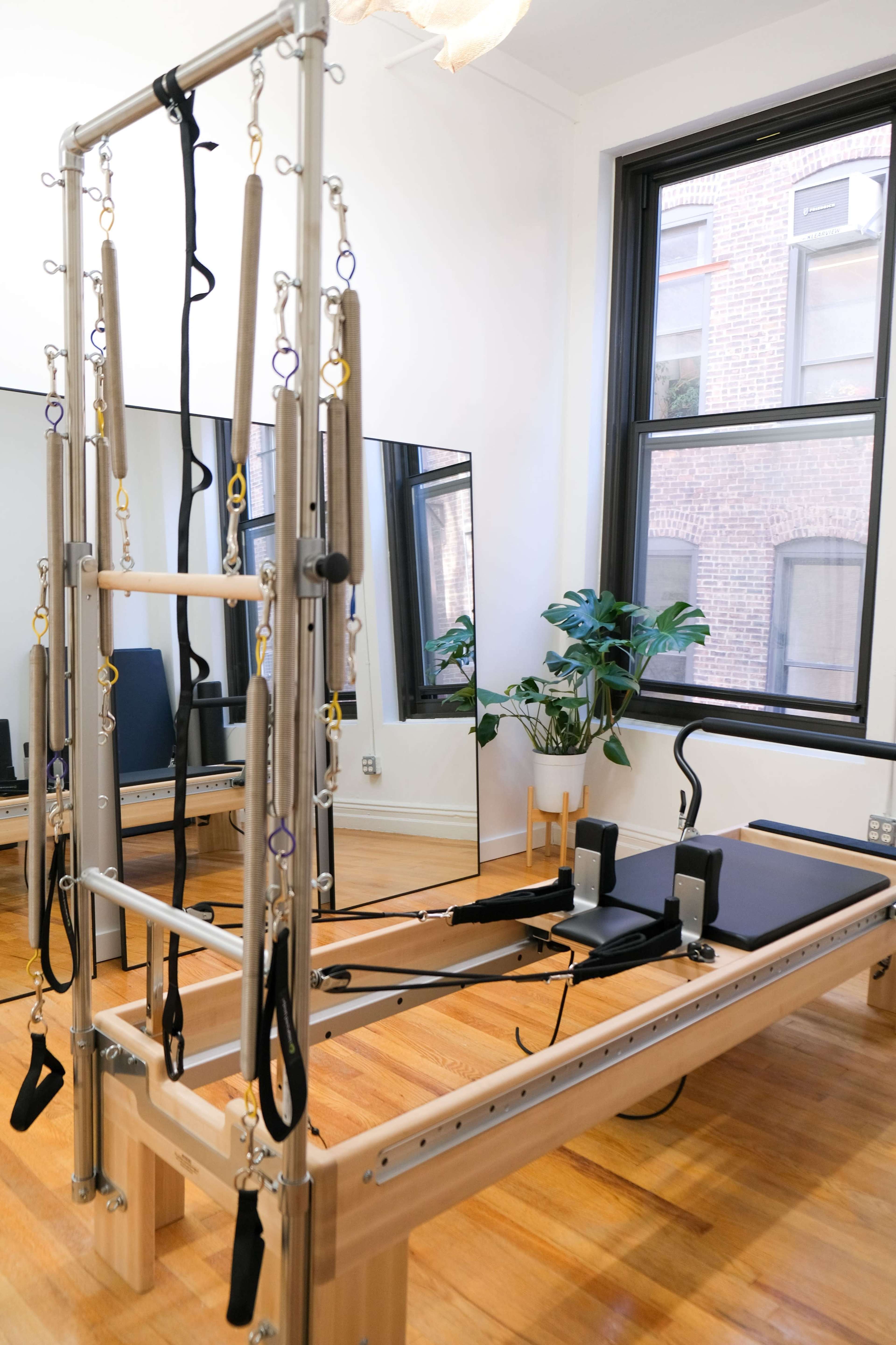 The image shows a Pilates reformer machine in a well-lit studio with large windows and a potted plant.