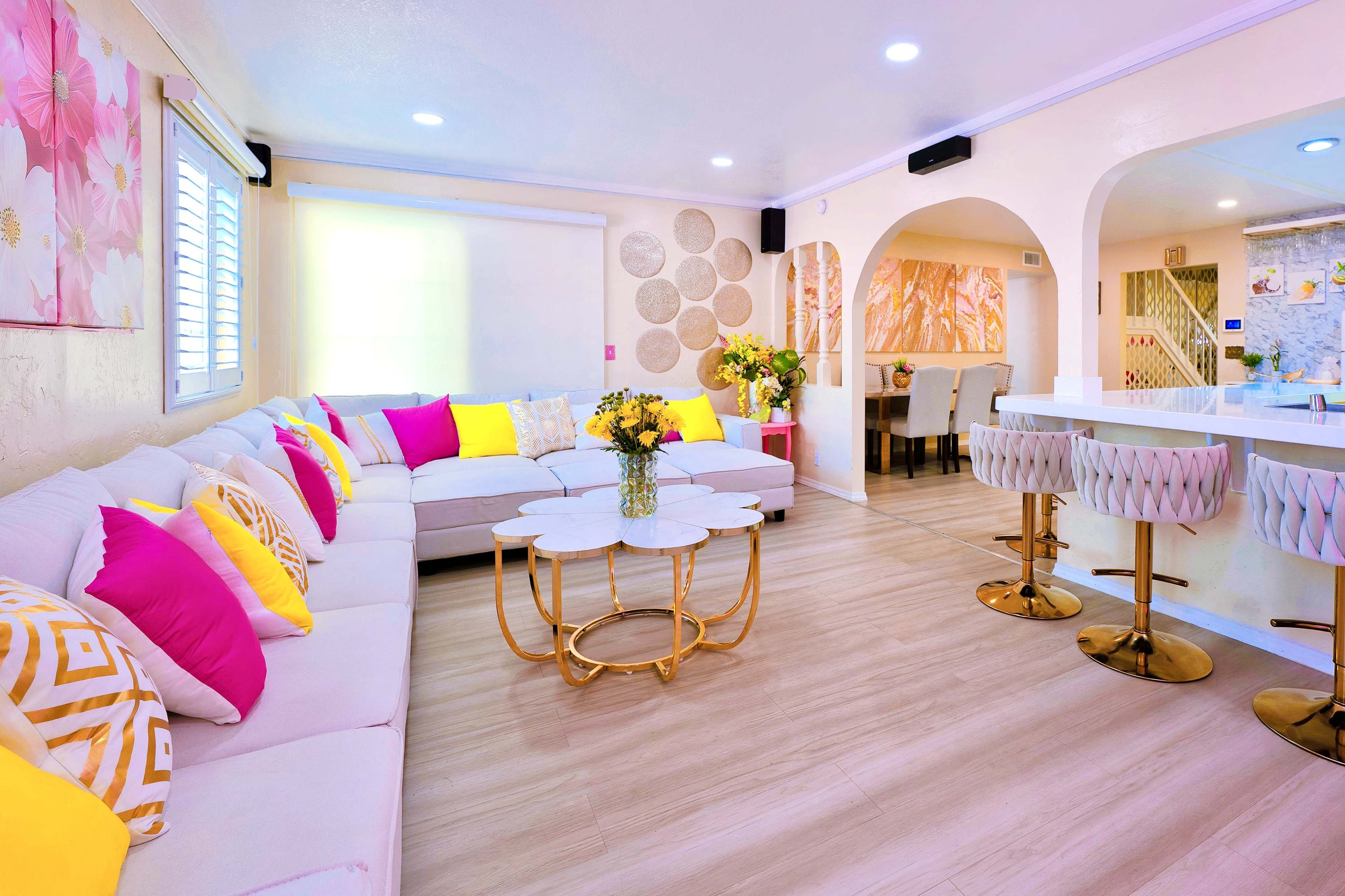 The image shows a bright, modern living area with a large sectional sofa adorned with pink and yellow pillows, a circular coffee table, and a dining area in the background.
