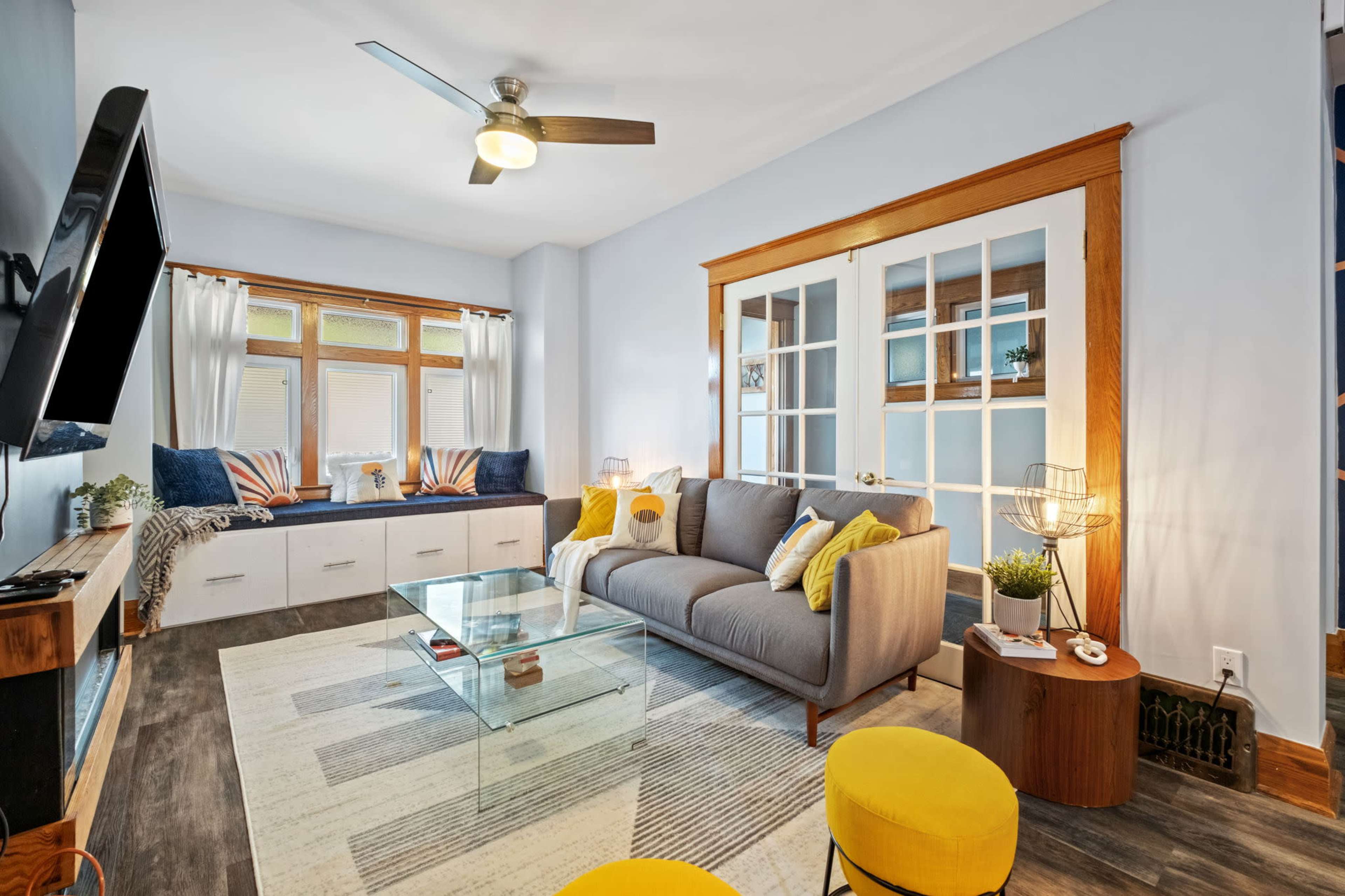 A modern living room features a gray sofa, yellow accent stools, a glass coffee table, and large windows with white curtains.