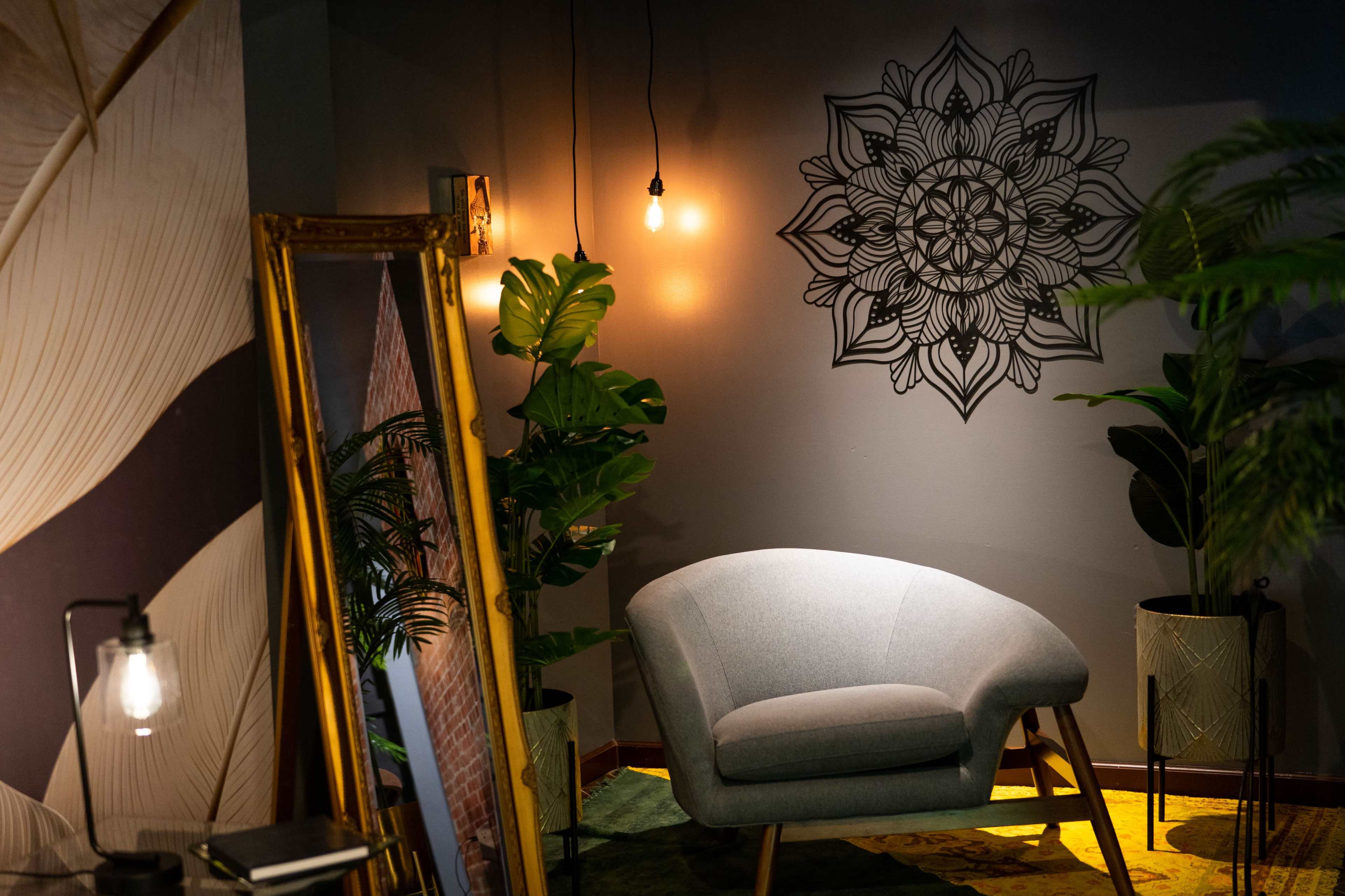 A cozy seating area with a gray chair next to a mirror, surrounded by tropical plants and adorned with a floral wall mural and pendant lights.
