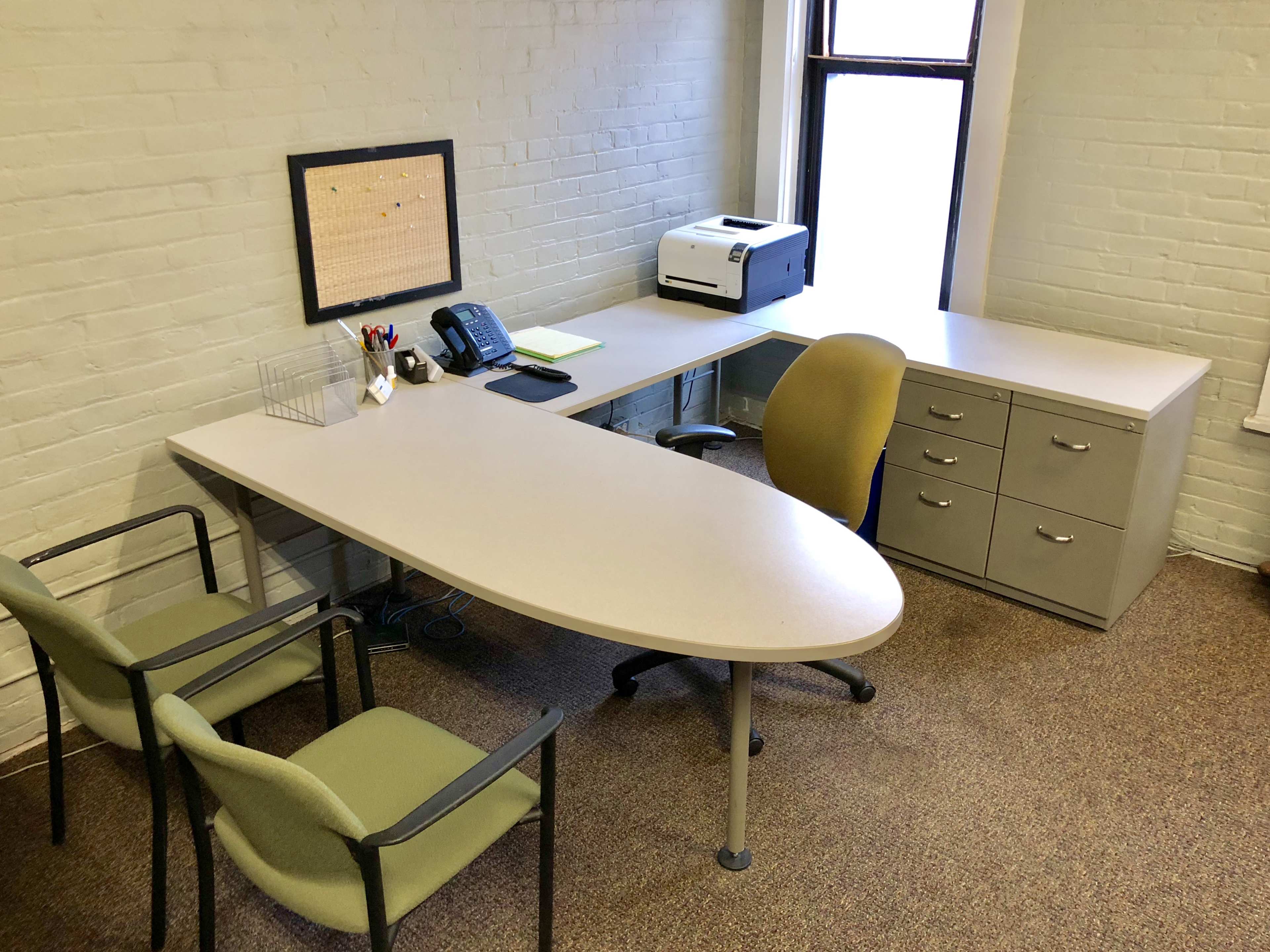 A small office space with a curved desk, a printer, a phone, two green chairs, and a filing cabinet against a light-colored brick wall.