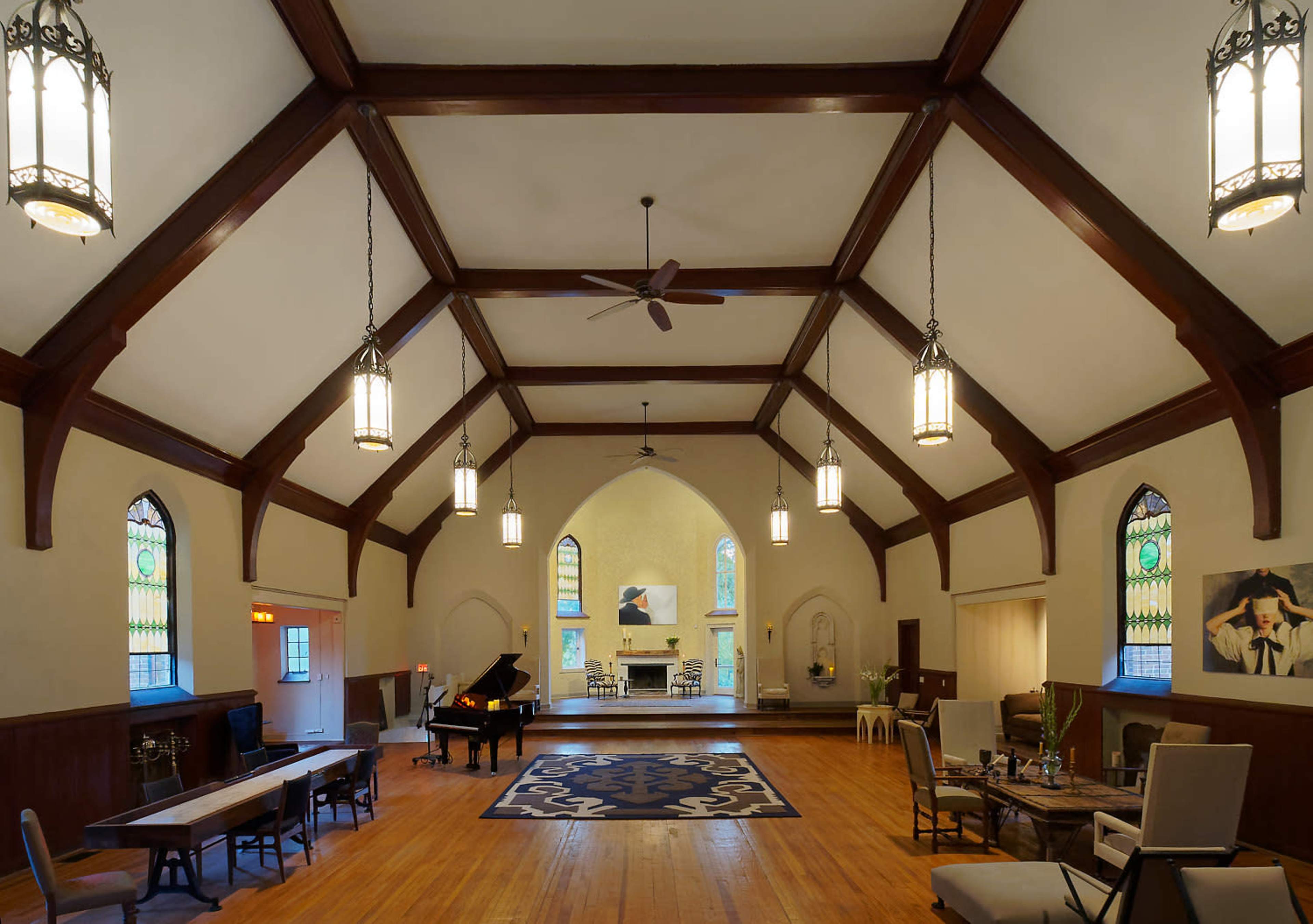 The interior of a spacious hall features wooden beams, large windows with stained glass, and a black piano positioned on a light-colored floor.