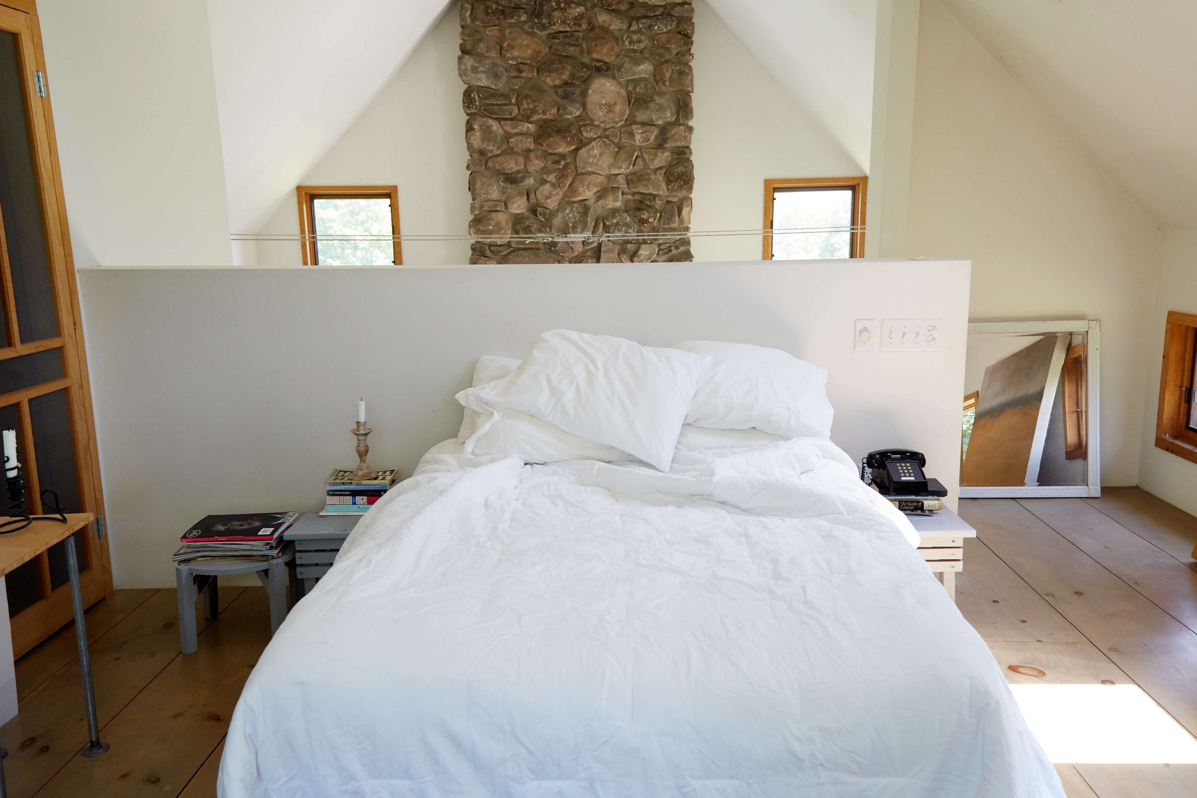A simple bedroom features a unmade bed with white bedding, a stone wall in the background, and a telephone on a nightstand.