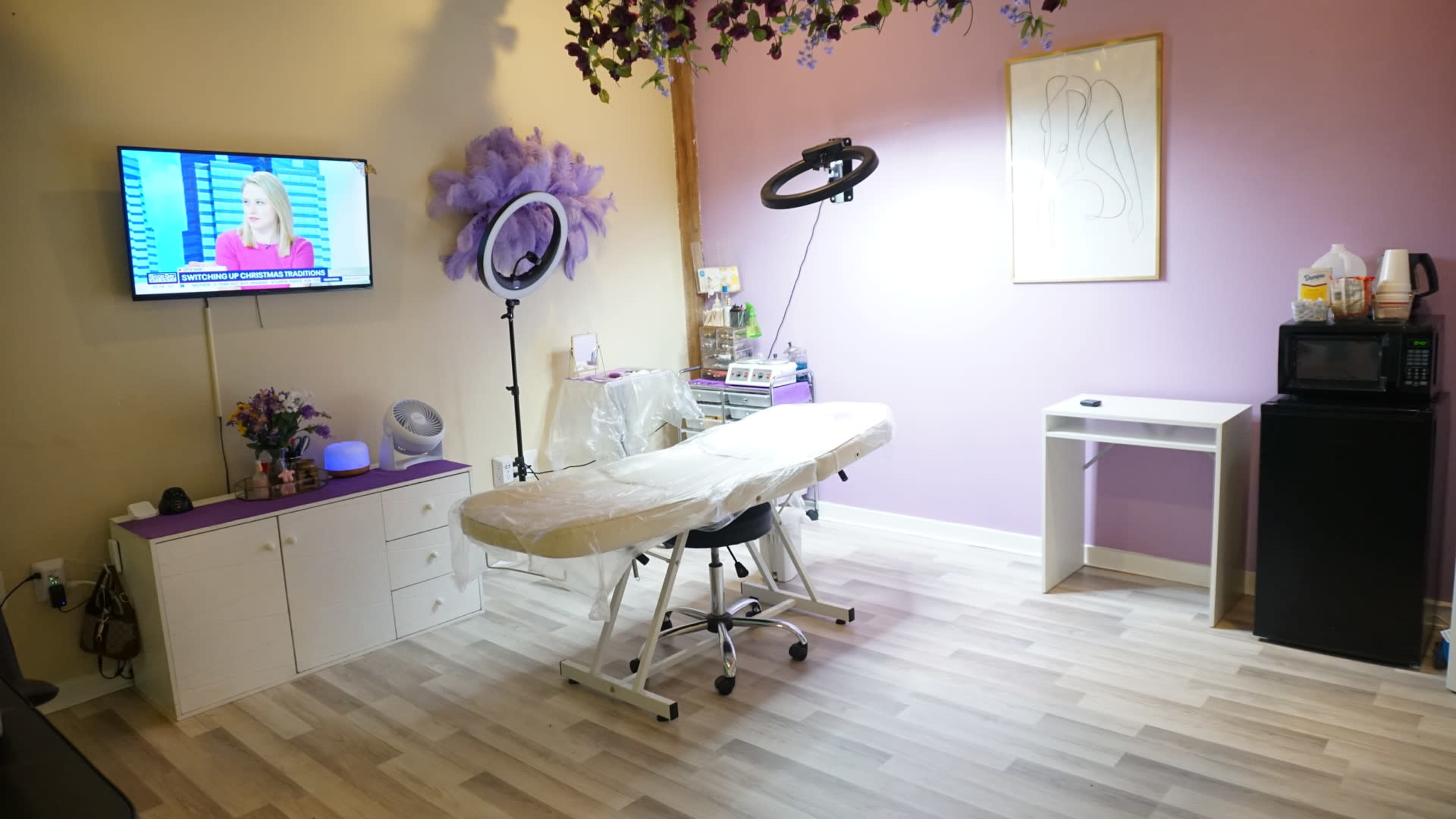 The image shows a small treatment room with a massage table, a television mounted on the wall, and various beauty equipment and furniture, including a microwave and a mini-fridge.