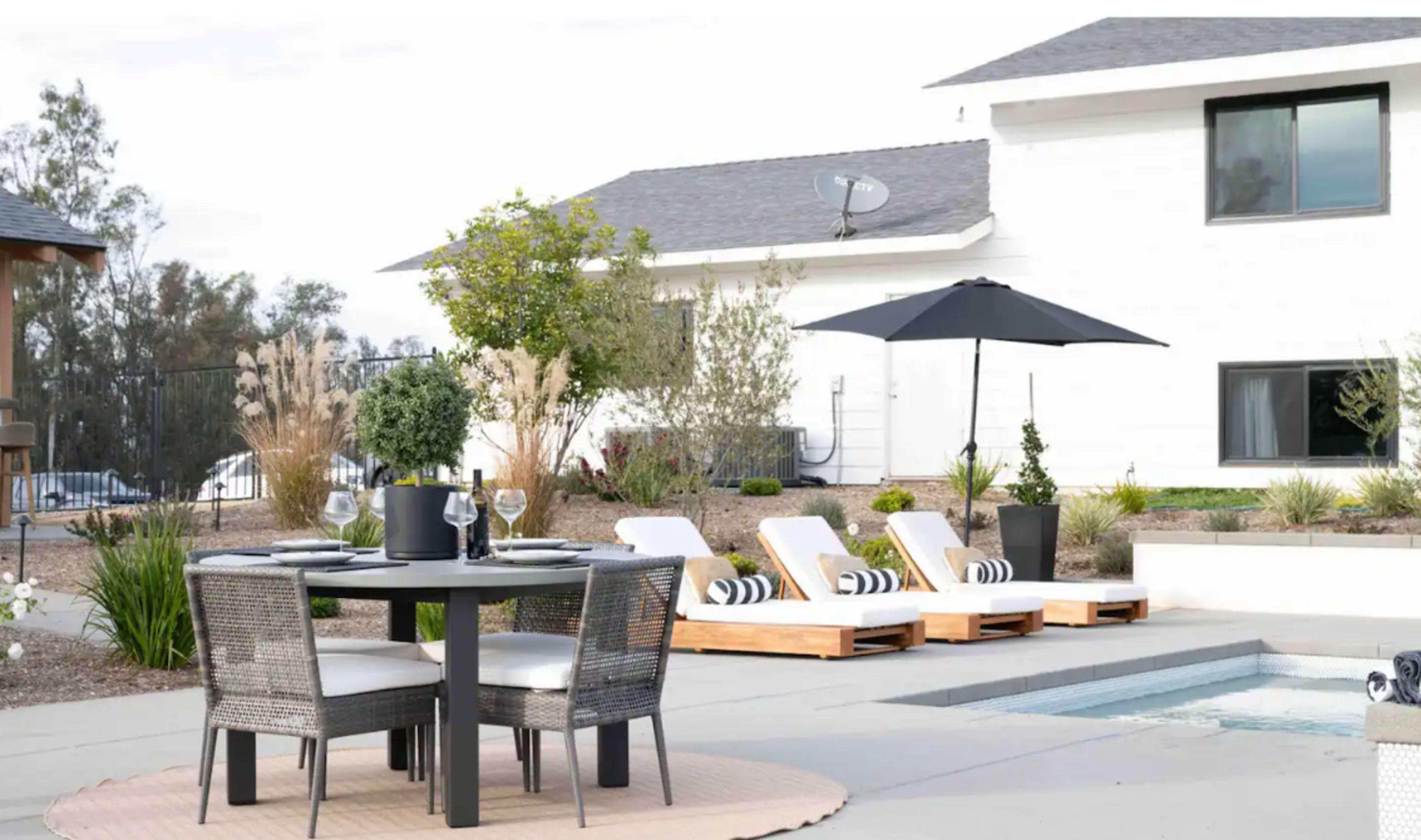 A patio features a round dining table with four chairs, a large umbrella, and two lounge chairs beside a small pool, set against a modern white house.