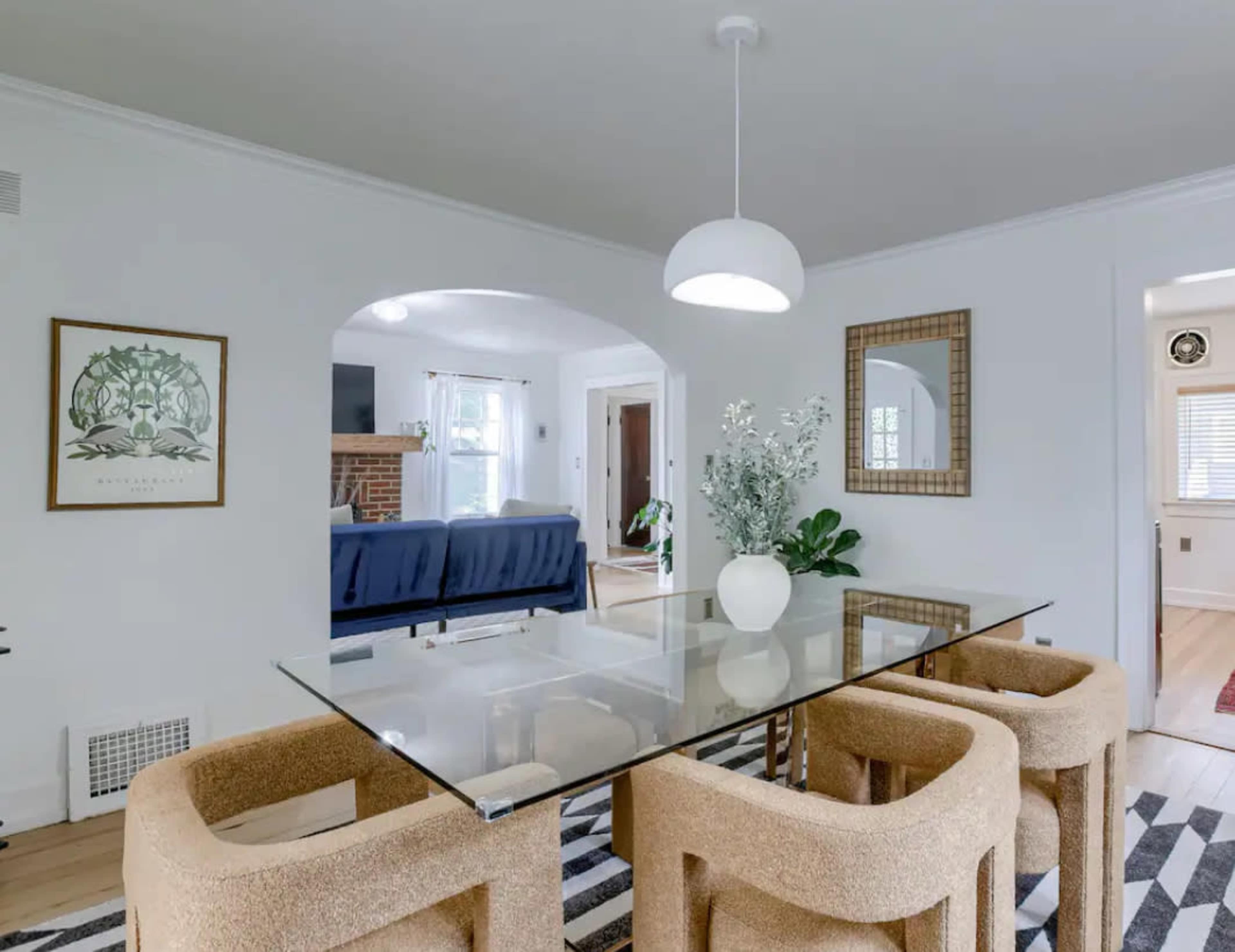 A dining area features a glass table surrounded by upholstered chairs, with a decorative plant and mirror in the backdrop.