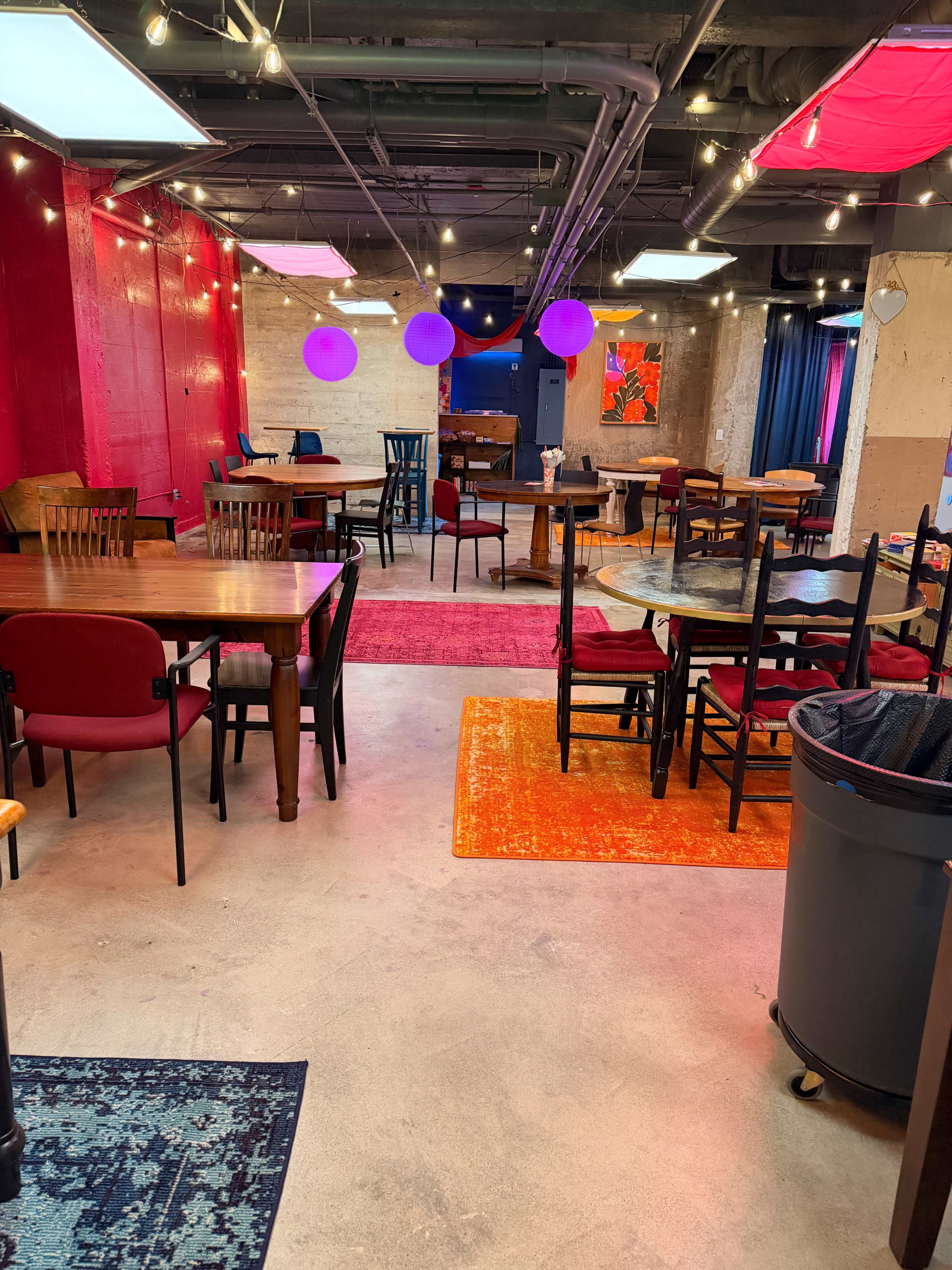 The image shows a spacious room filled with various tables and chairs, colorful rugs on the floor, and decorative lighting overhead.