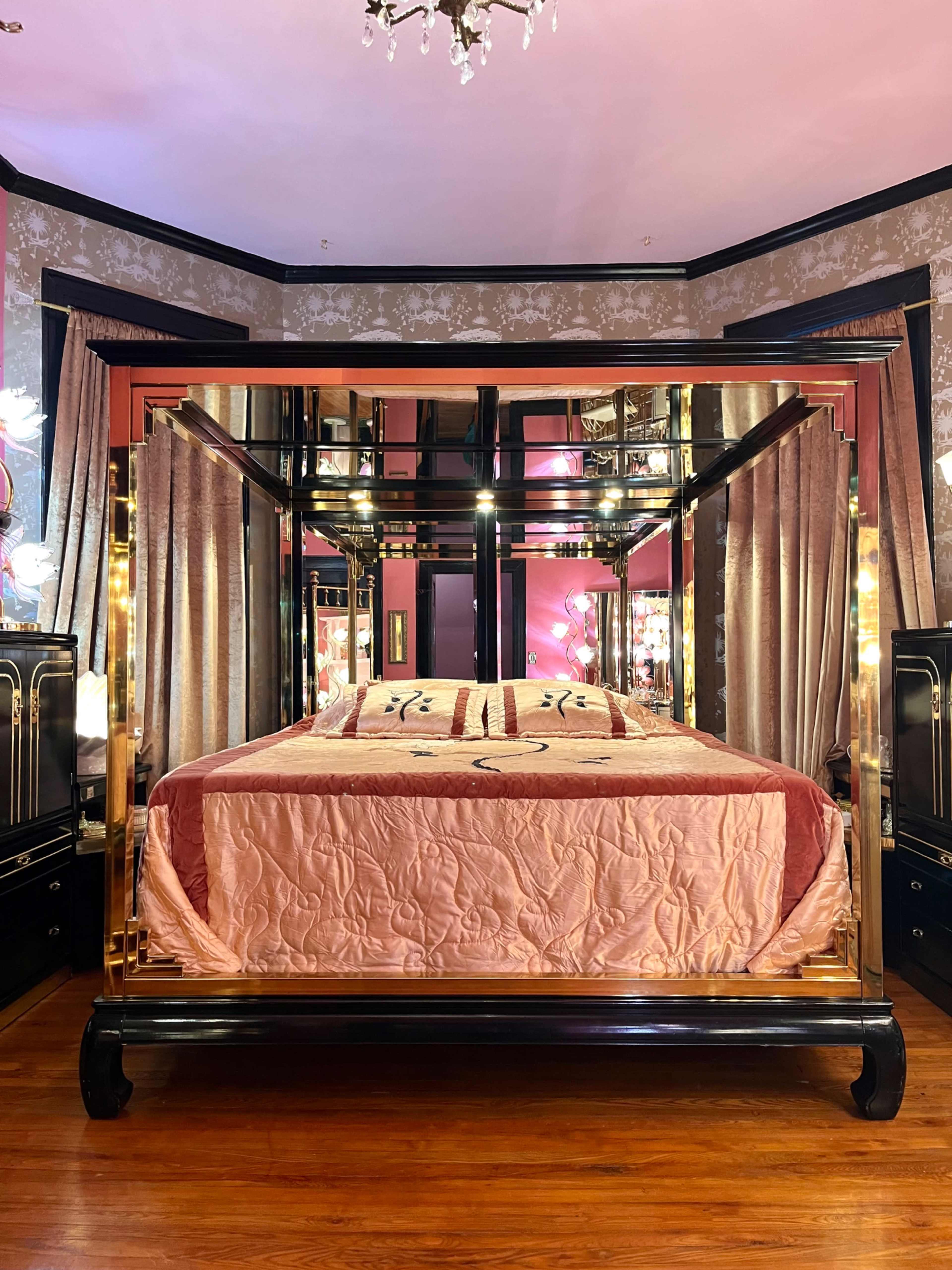 The image shows a bedroom featuring a large, ornate four-poster bed with a pink quilt and mirrored accents, surrounded by dark cabinetry and stylish decor.