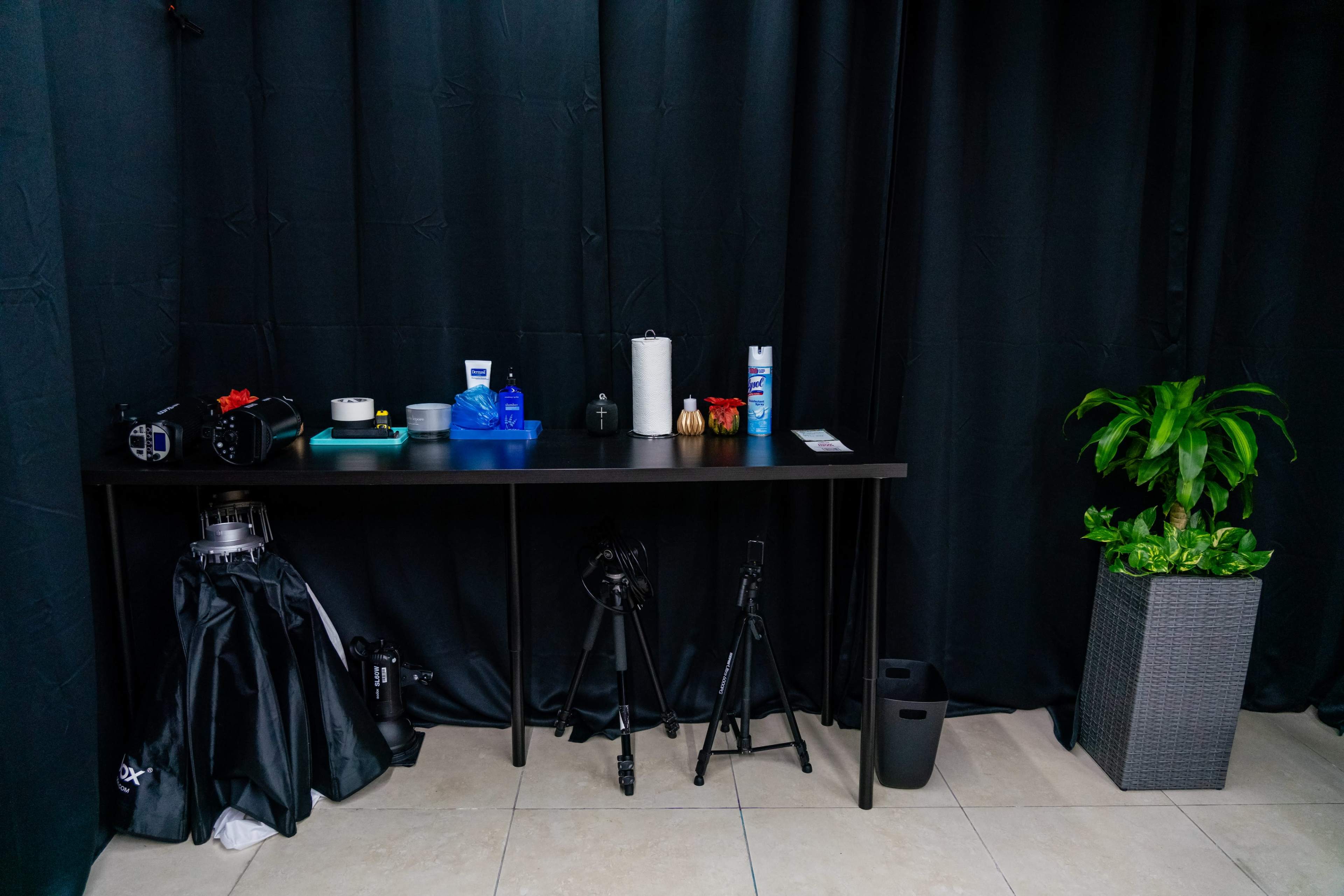 A black table is positioned against a dark curtain and is arranged with various items, including cameras, cleaning supplies, and a potted plant.