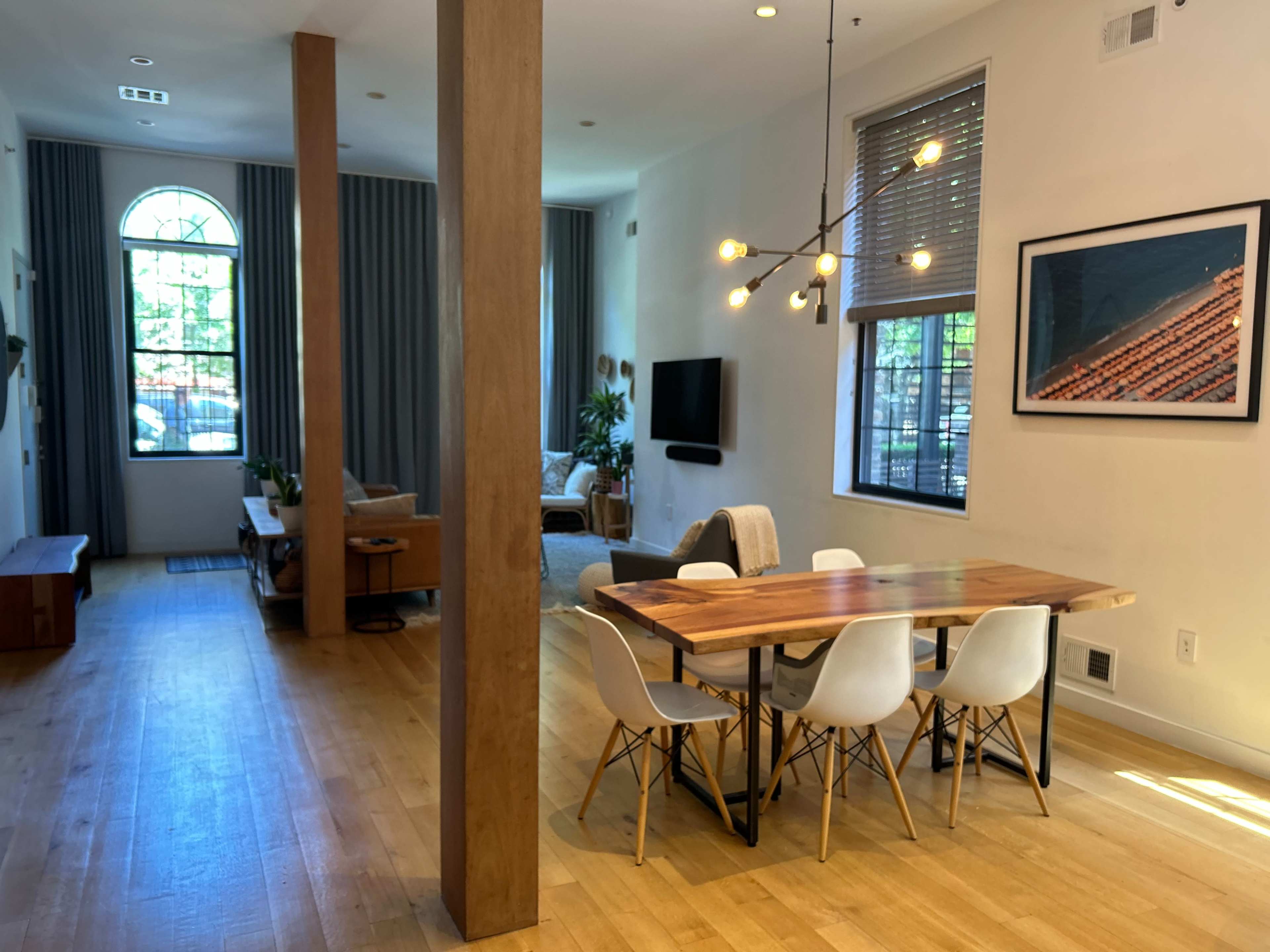 A modern open-concept living space featuring a dining area with a wooden table and white chairs, a living area with a sofa, and large windows providing natural light.