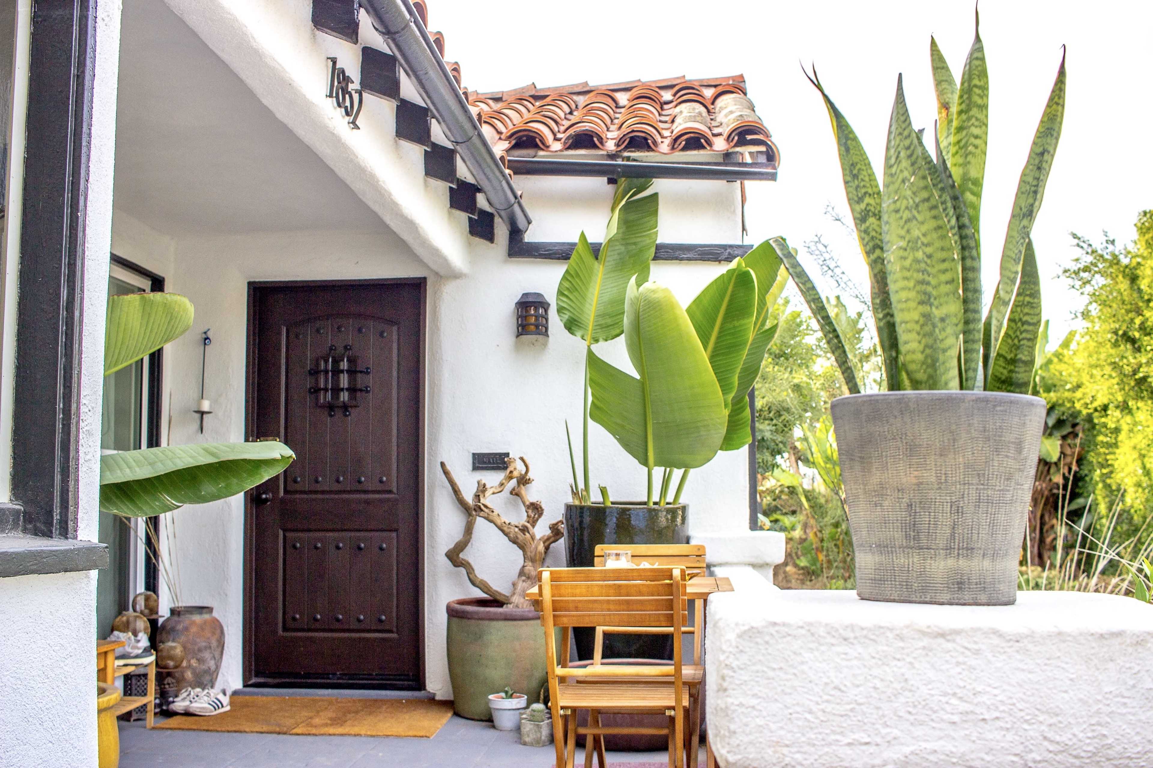 A house entrance featuring a wooden door, potted plants, and a small wooden table with chairs on a paved area.