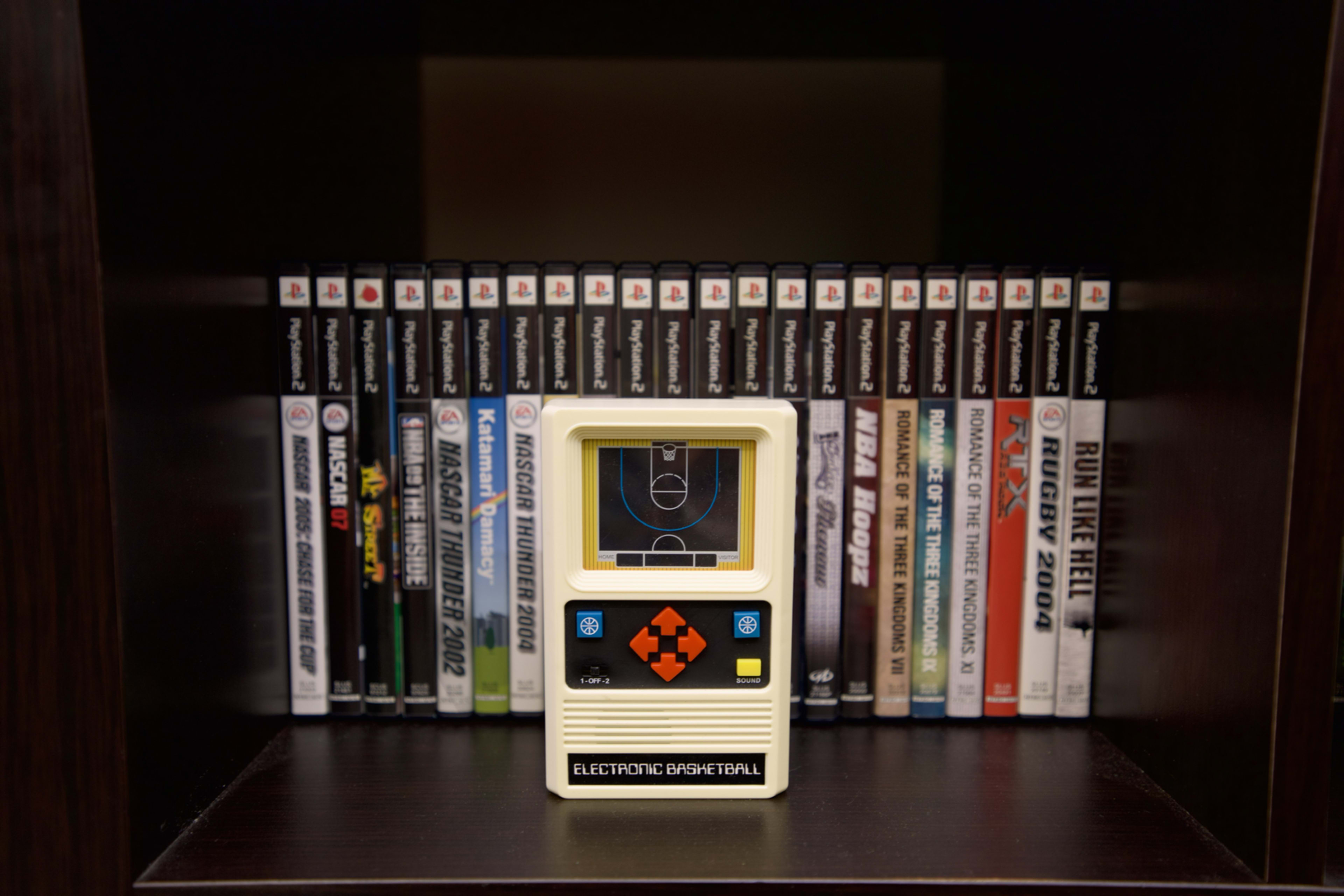 An electronic basketball handheld game is displayed in front of a shelf filled with various video game cases.