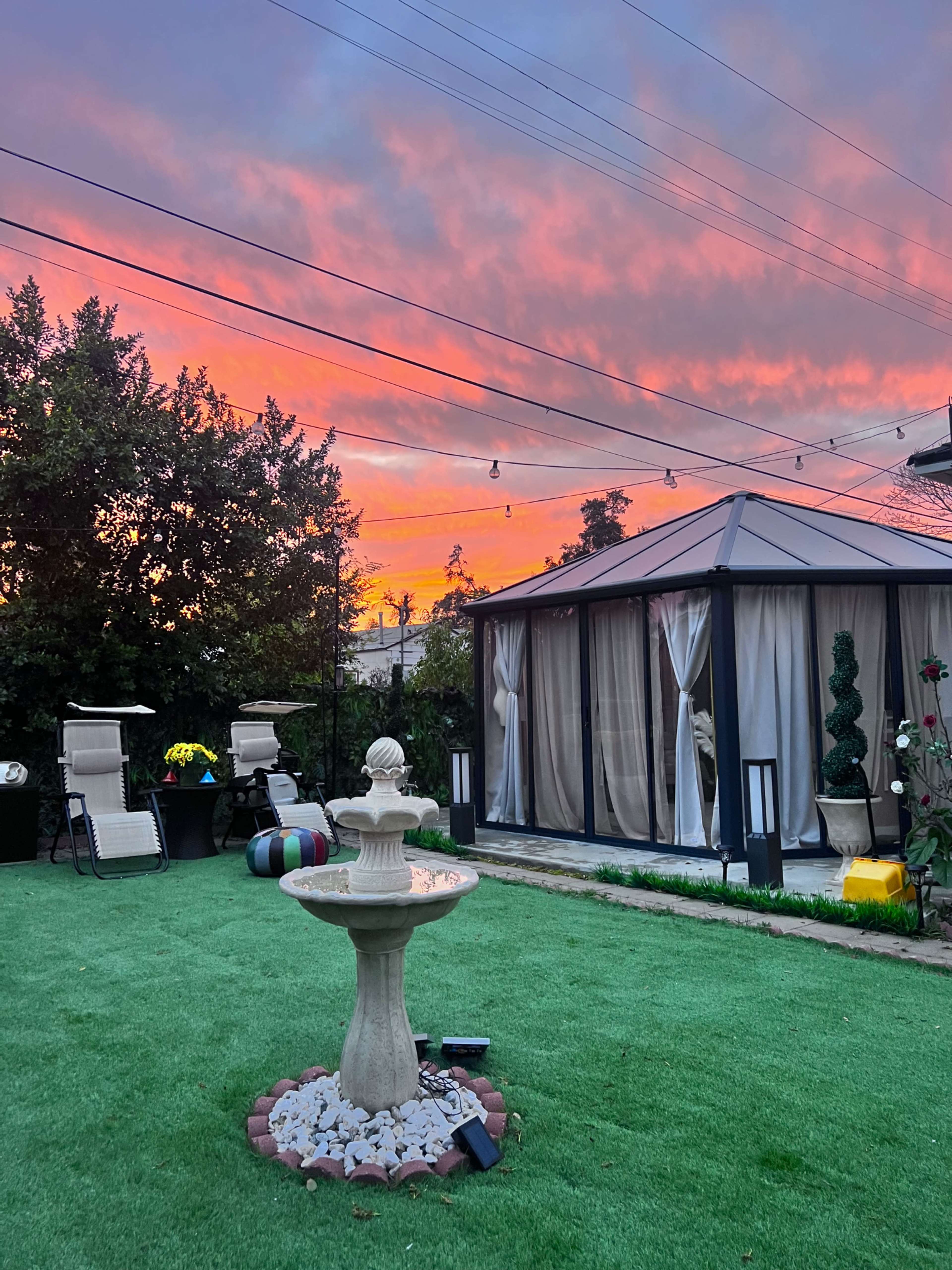 The scene features a landscaped backyard with a fountain in the center, surrounded by chairs and a gazebo, under a colorful sunset sky.