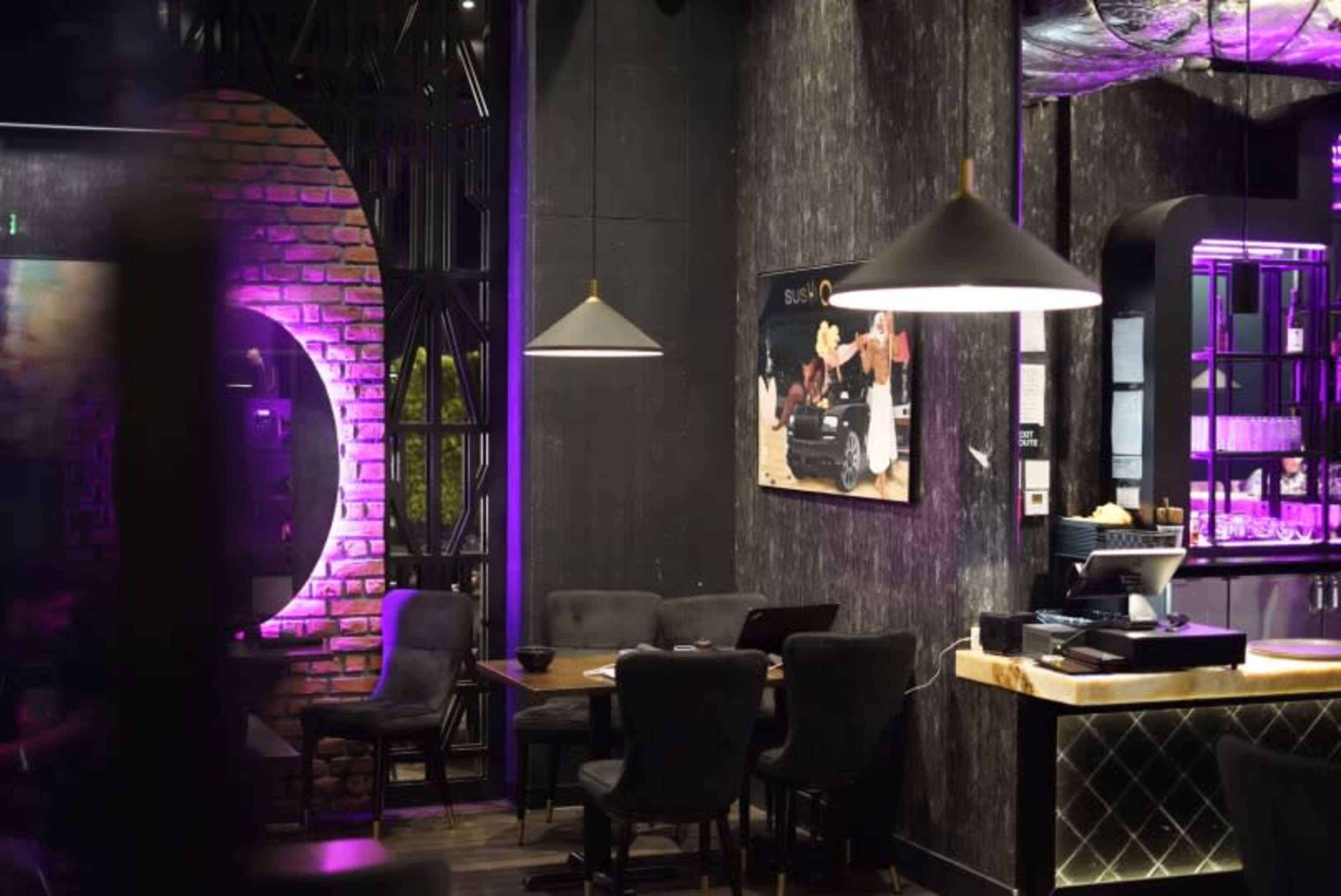 The image shows a modern restaurant interior featuring dark walls, purple lighting, and a combination of wooden and upholstered furniture.