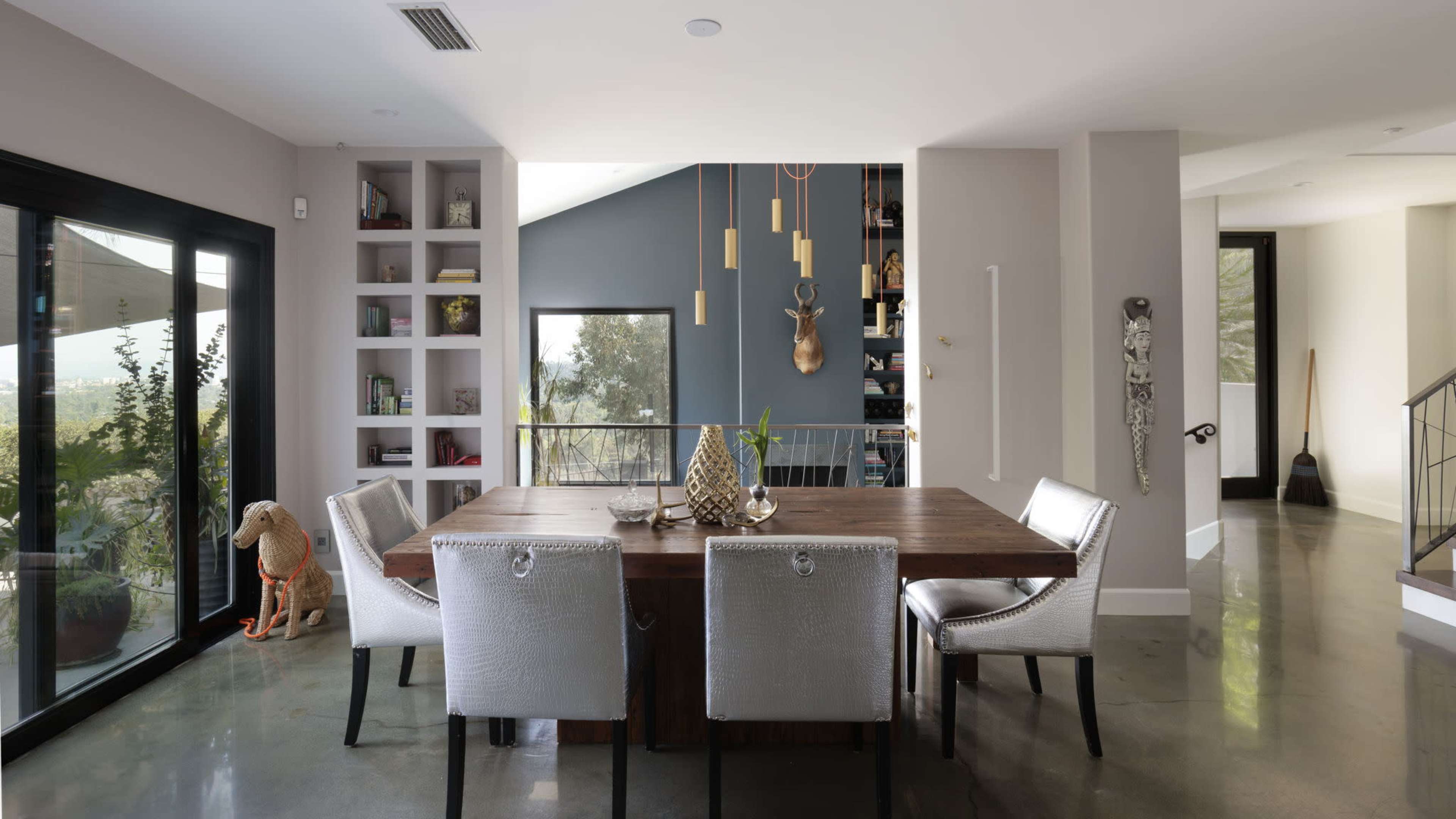 The image shows a modern dining area with a wooden table surrounded by four metallic chairs, shelves with decorative items, and a large glass door leading to an outdoor space.