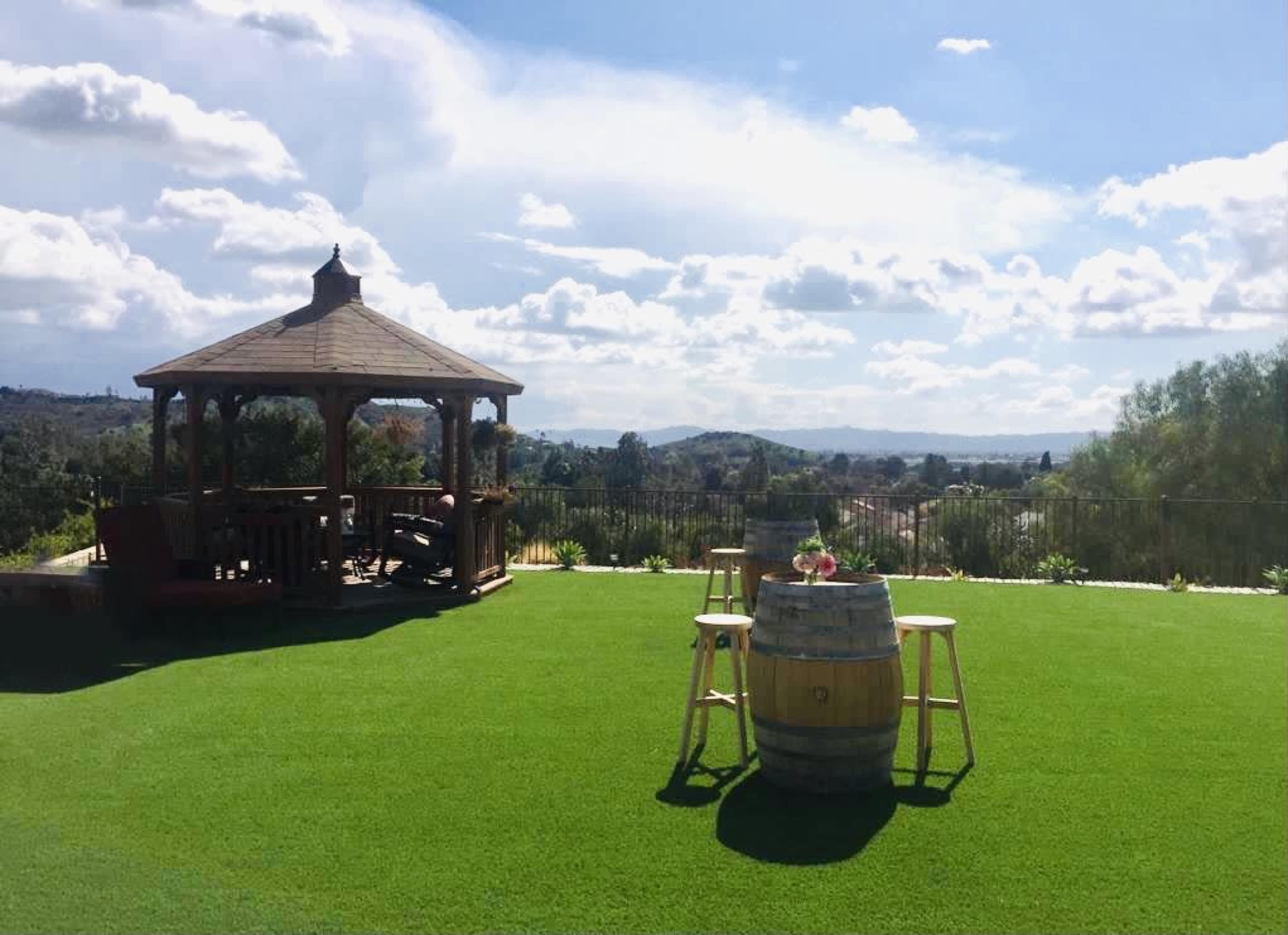 A gazebo stands on a green lawn overlooking rolling hills under a partly cloudy sky, with a wooden barrel and three stools nearby.