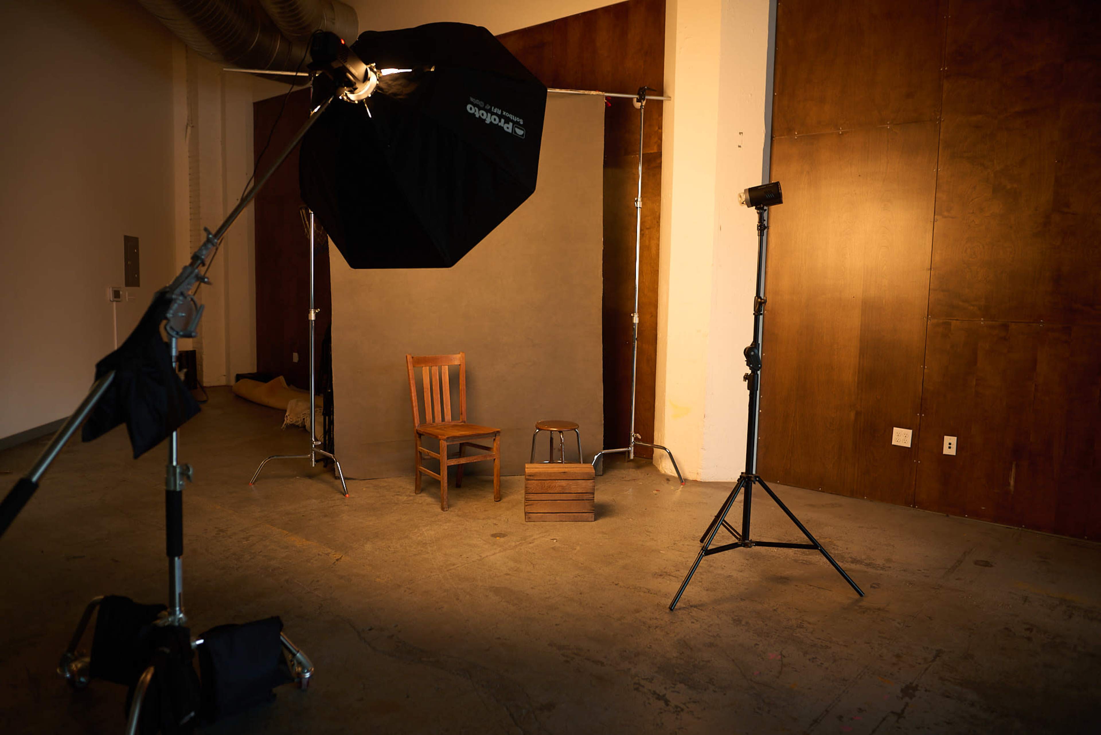 The image shows a photography studio setup with a light source, a black umbrella softbox, a wooden chair, a small stool, and a backdrop against a wooden wall.