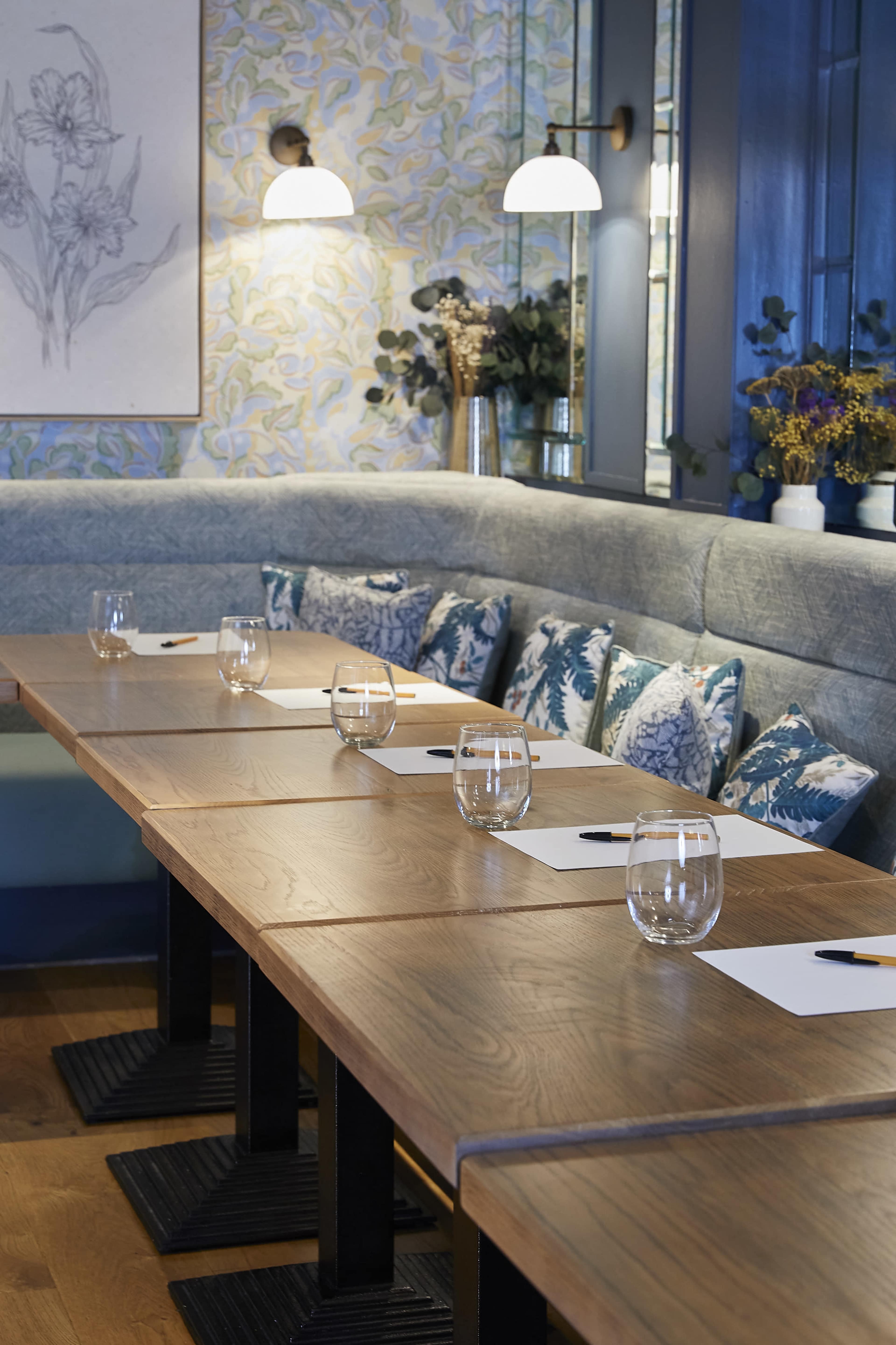The image shows a restaurant interior featuring a long wooden table set with glasses and paper notepads, surrounded by upholstered benches with decorative pillows against a floral wallpaper.