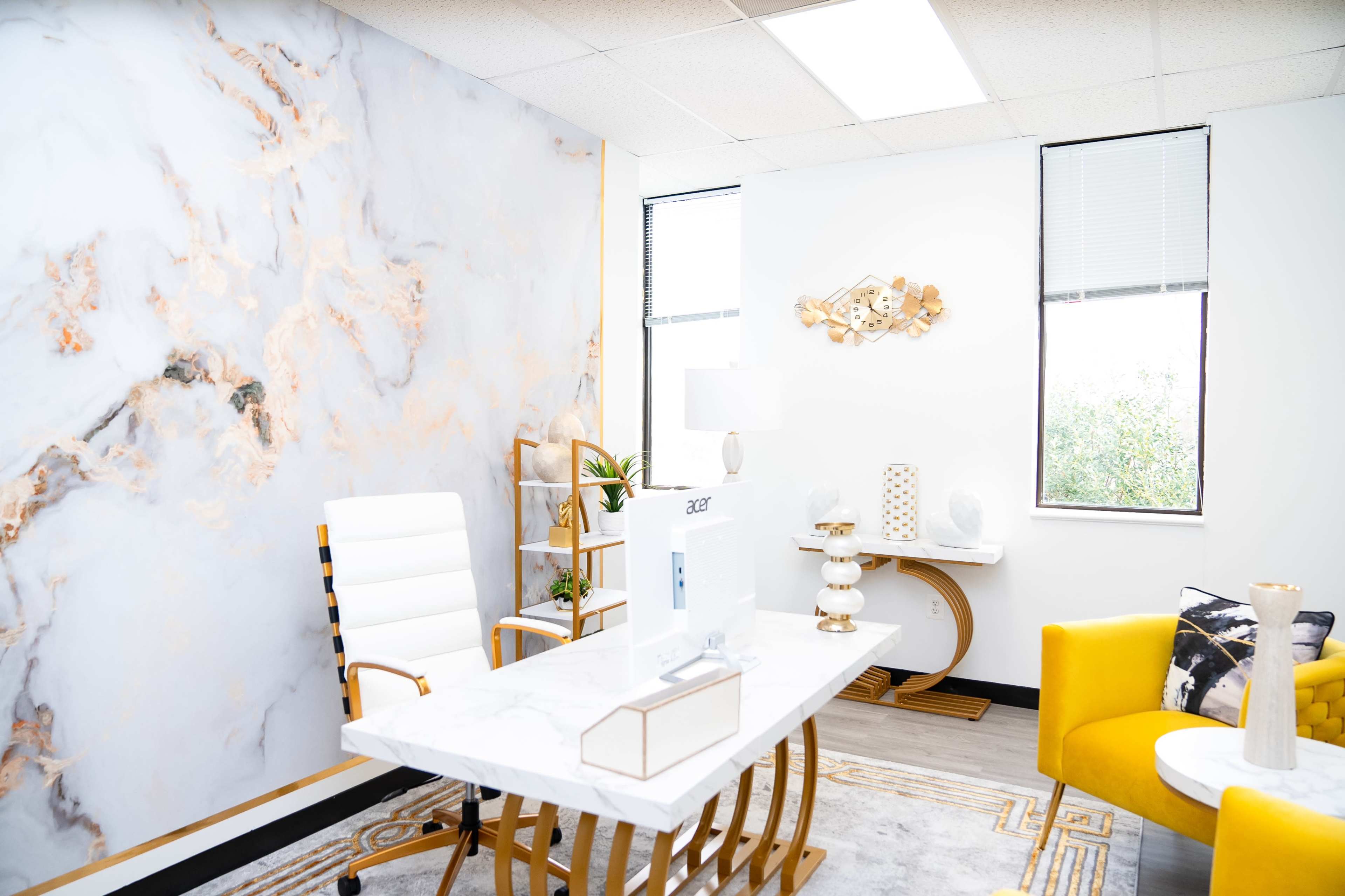 A modern office space features a marble-patterned accent wall, a white desk with gold legs, a yellow chair, and large windows allowing natural light.
