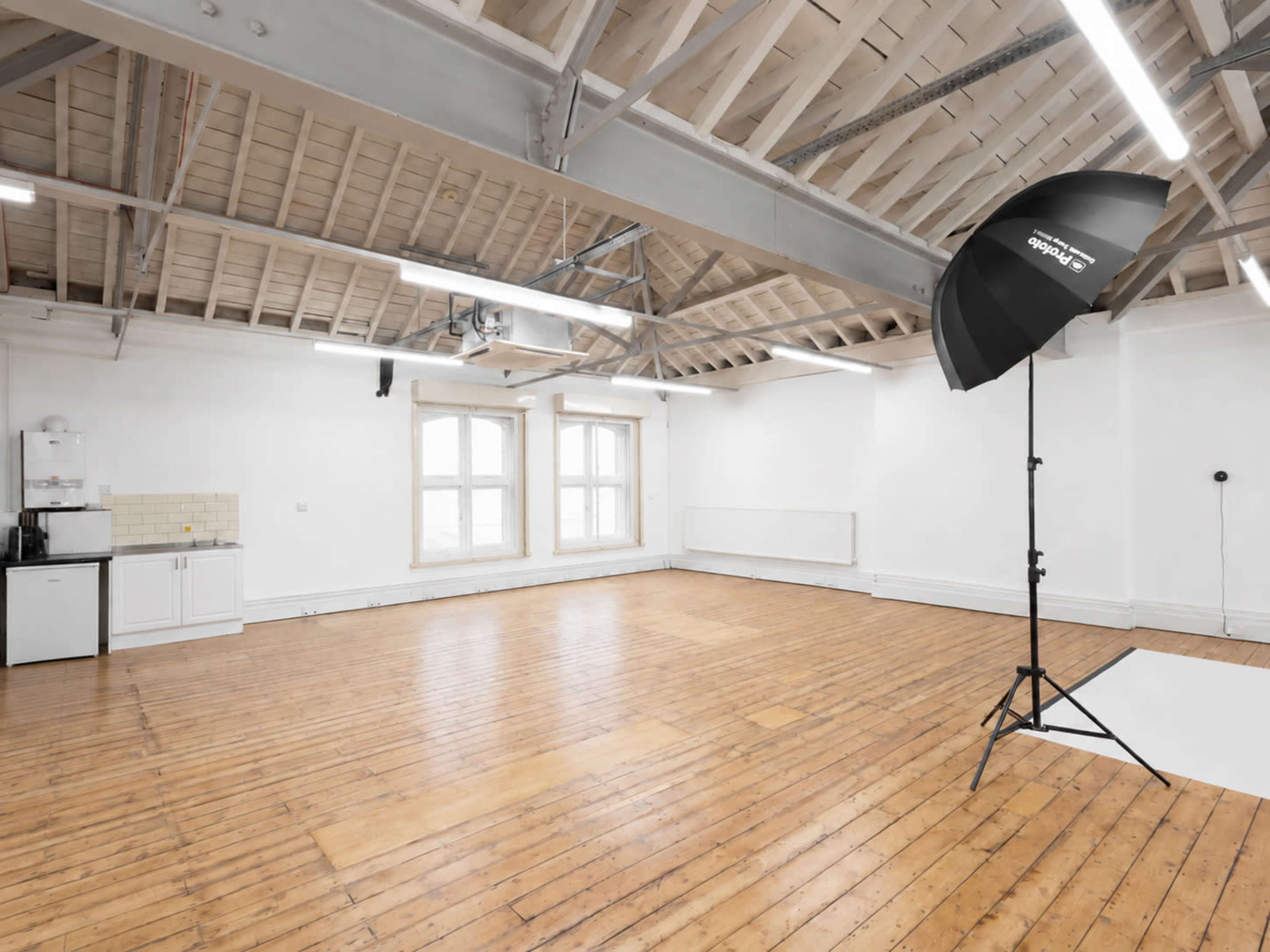 The image shows an empty studio space with wooden floors, white walls, and a photography lighting setup in one corner.