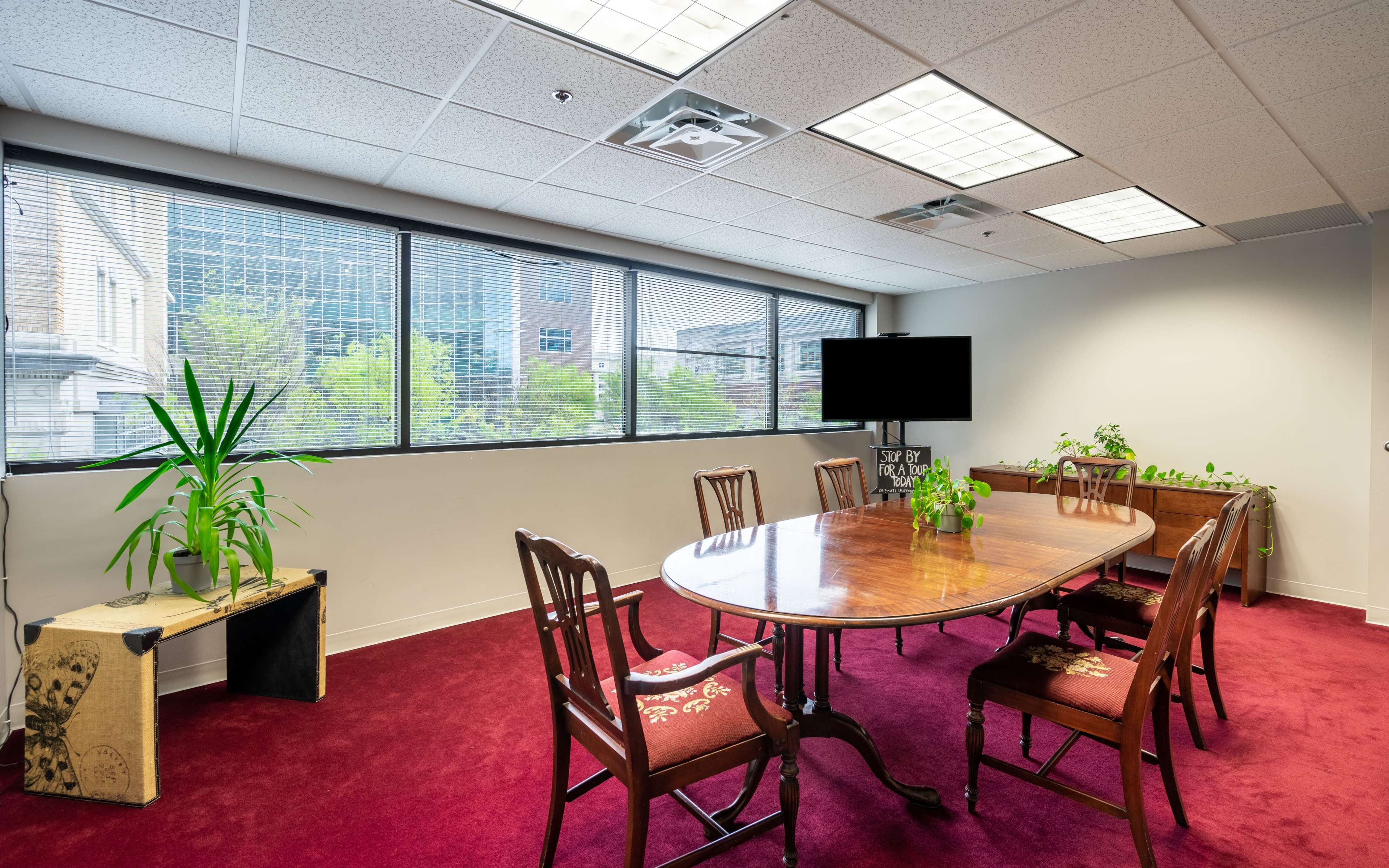 A conference room features a long wooden table surrounded by chairs, with large windows providing natural light and plants placed around the space.
