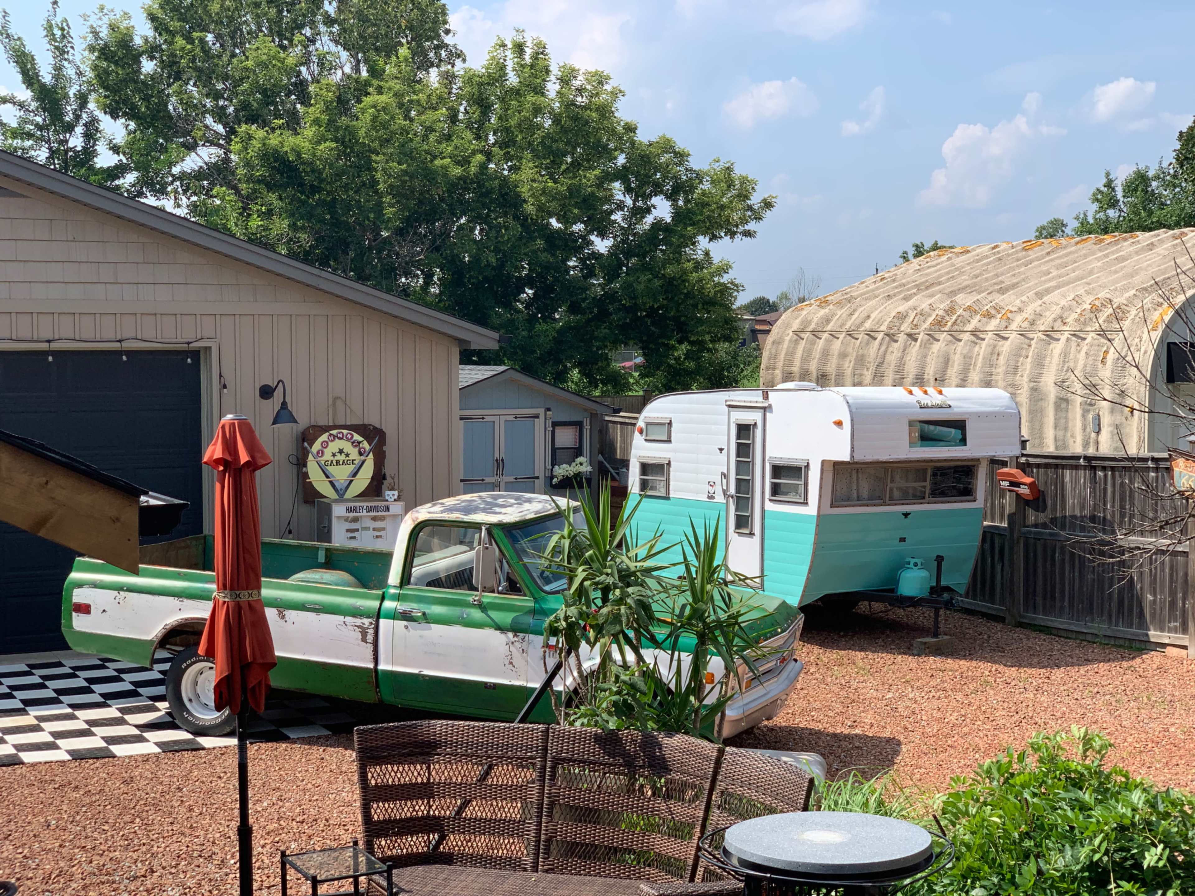 The scene features a vintage pickup truck parked beside a turquoise and white camper in a gravel yard with a checkered patio.
