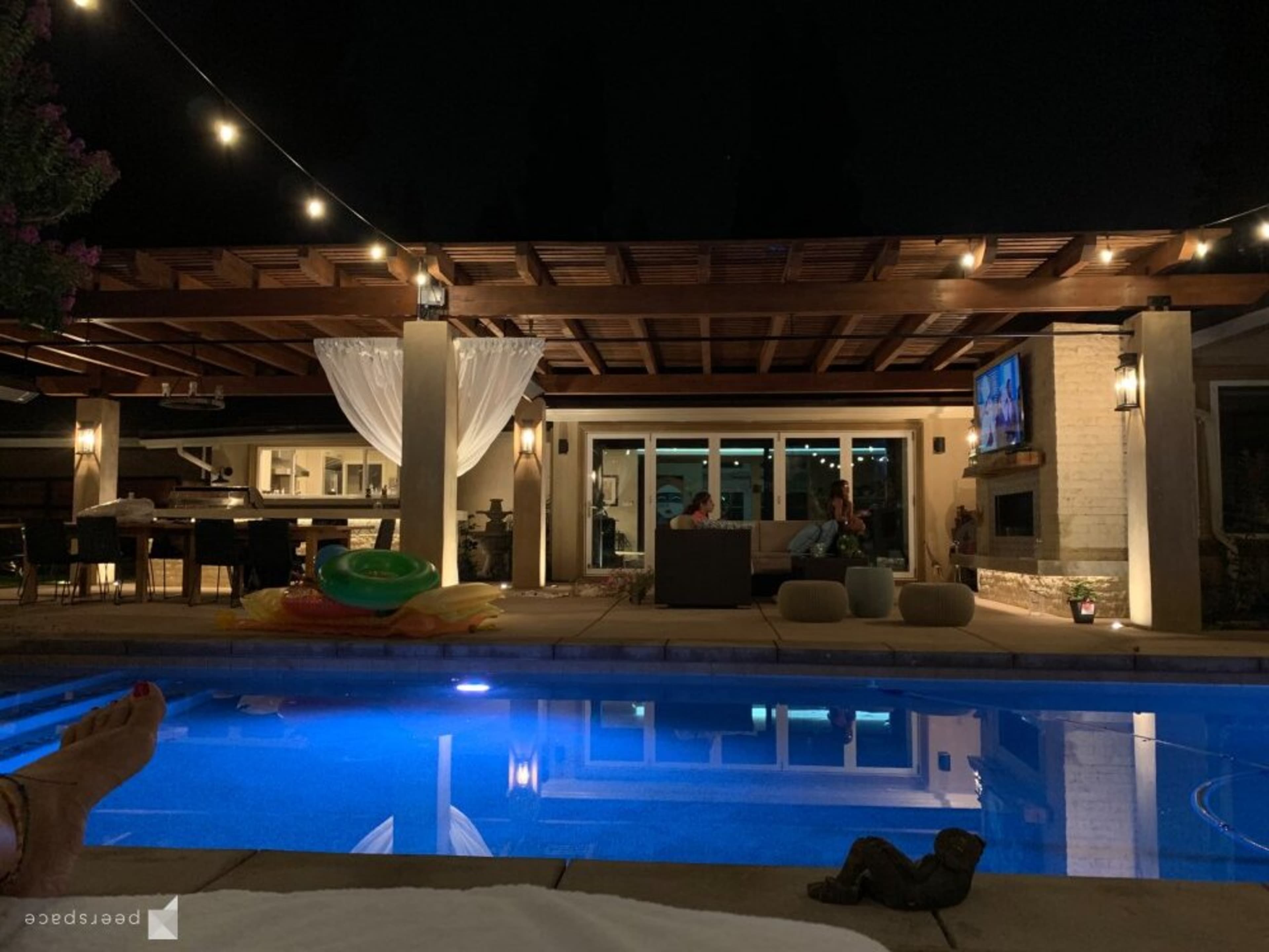 A well-lit outdoor space features a swimming pool reflecting lights, a seating area, and a wooden pergola adorned with string lights at night.