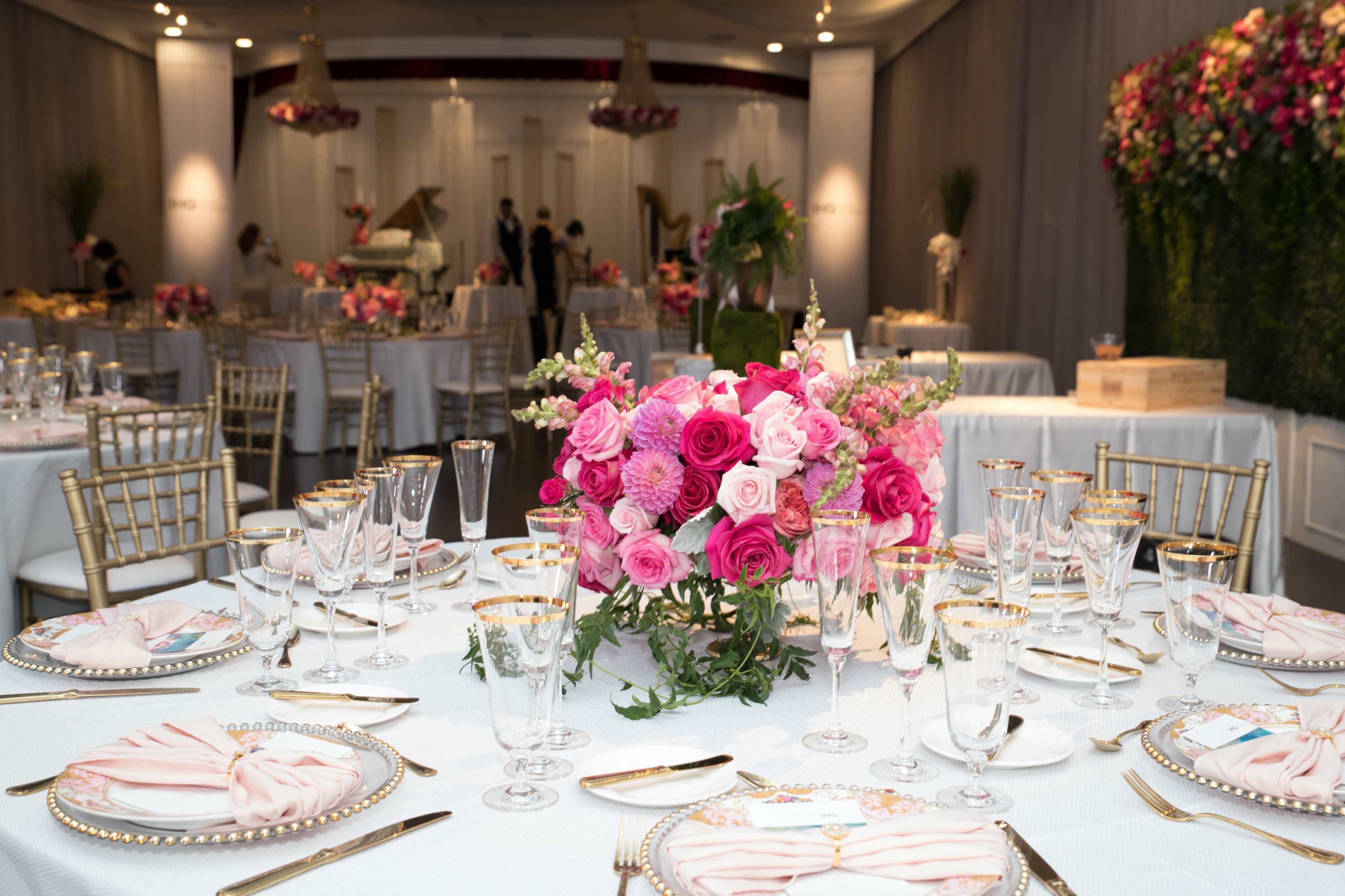 A elegantly set dining area features round tables adorned with floral centerpieces, fine china, and crystal glassware, with a backdrop of draped fabric and floral arrangements.