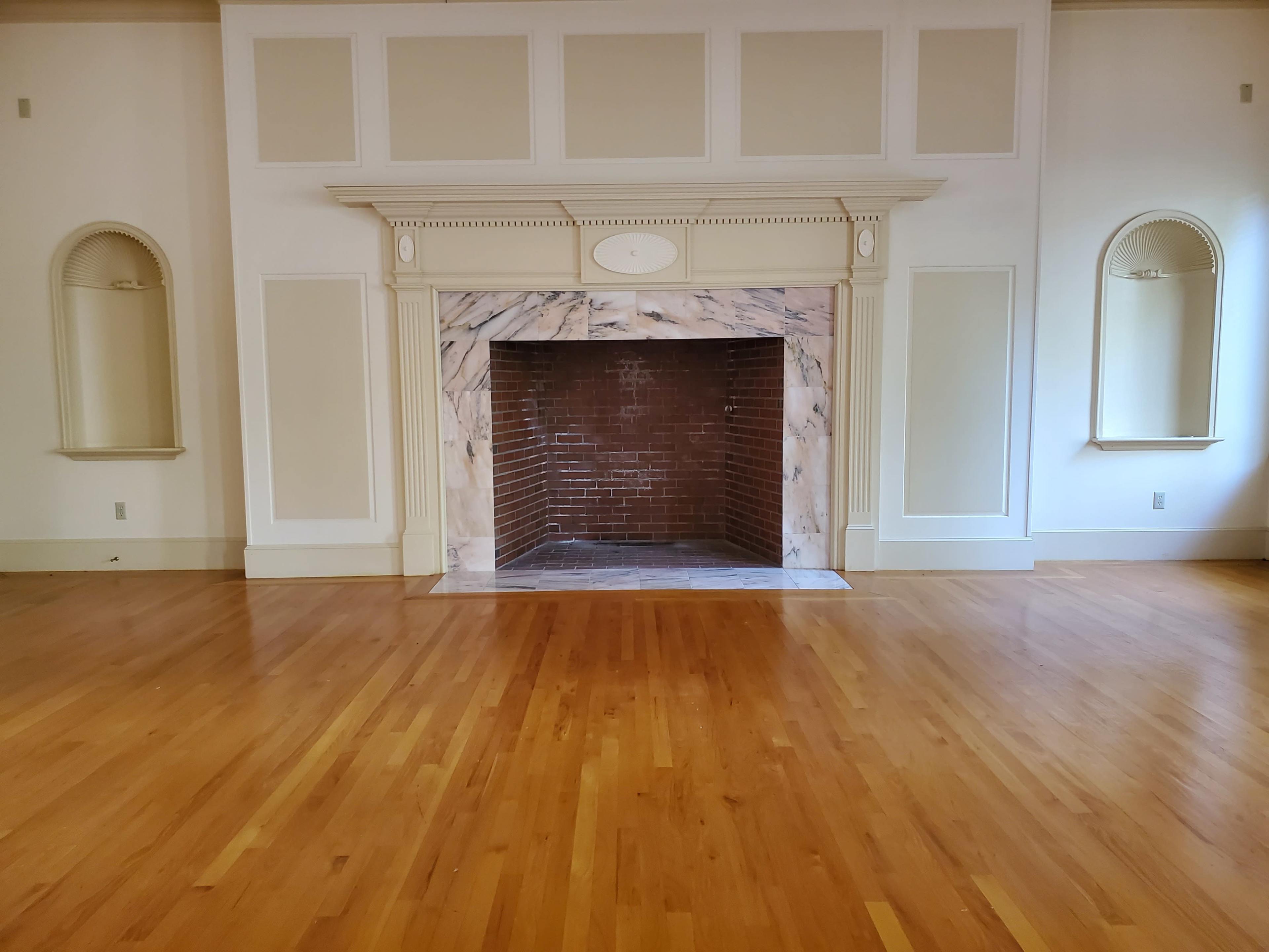 A room with a large fireplace made of marble, flanked by two small alcoves, and has polished wooden flooring.