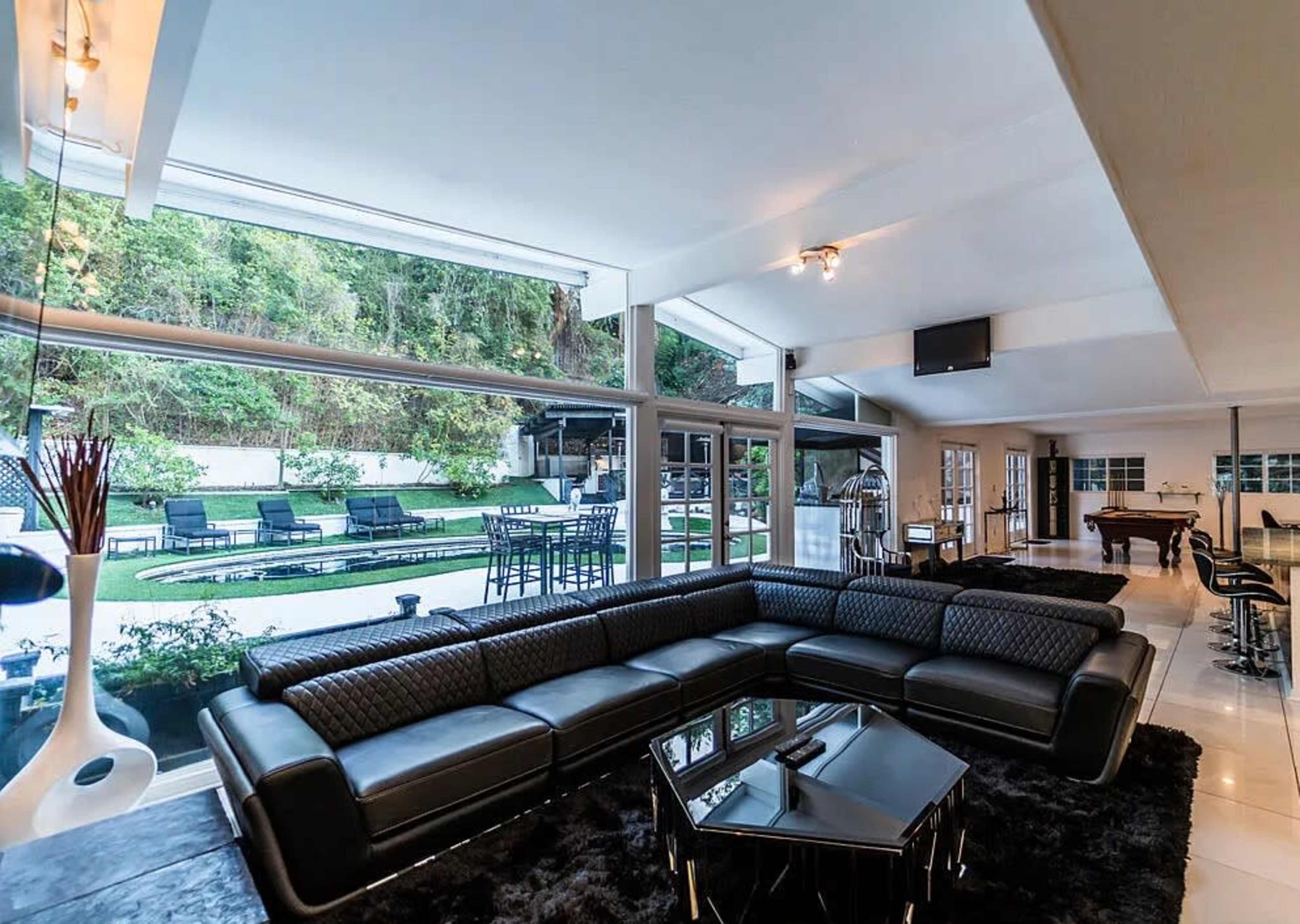 The image shows a modern living room with a black sectional sofa, large windows overlooking a backyard with a patio and swimming pool, and a grand piano in the background.