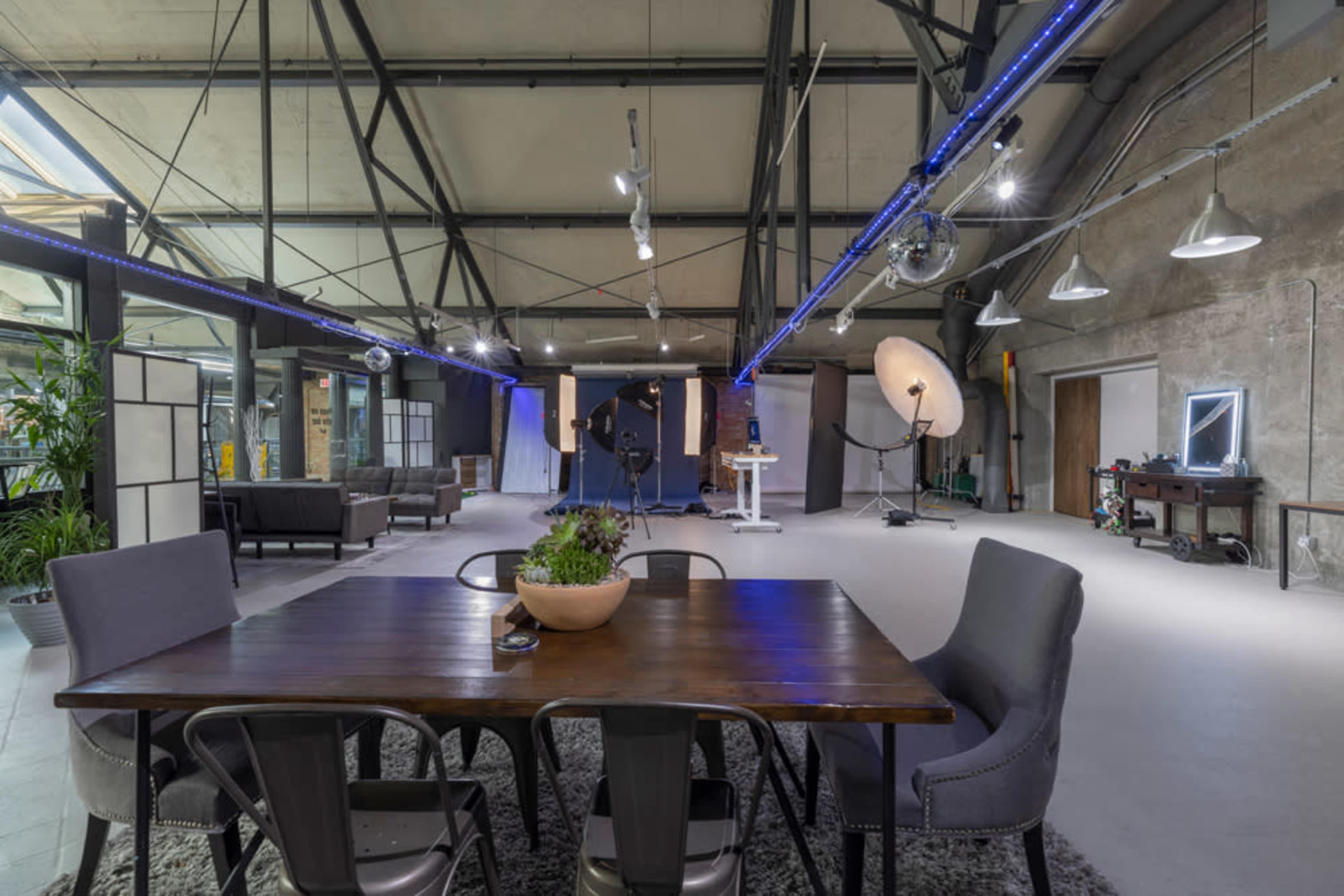 A spacious industrial-style studio with high ceilings, a wooden dining table, modern chairs, and photography equipment in the background.