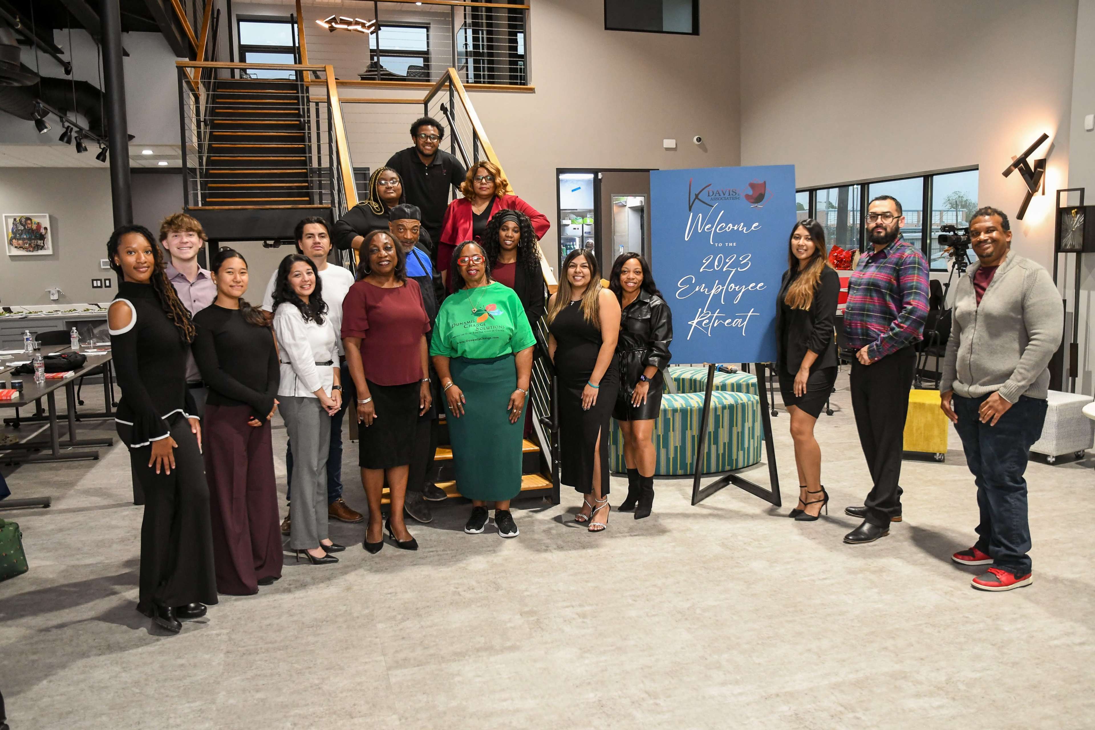 A group of diverse individuals stands together at an indoor venue, posing for a photo in front of a welcome sign for a 2023 employee retreat.