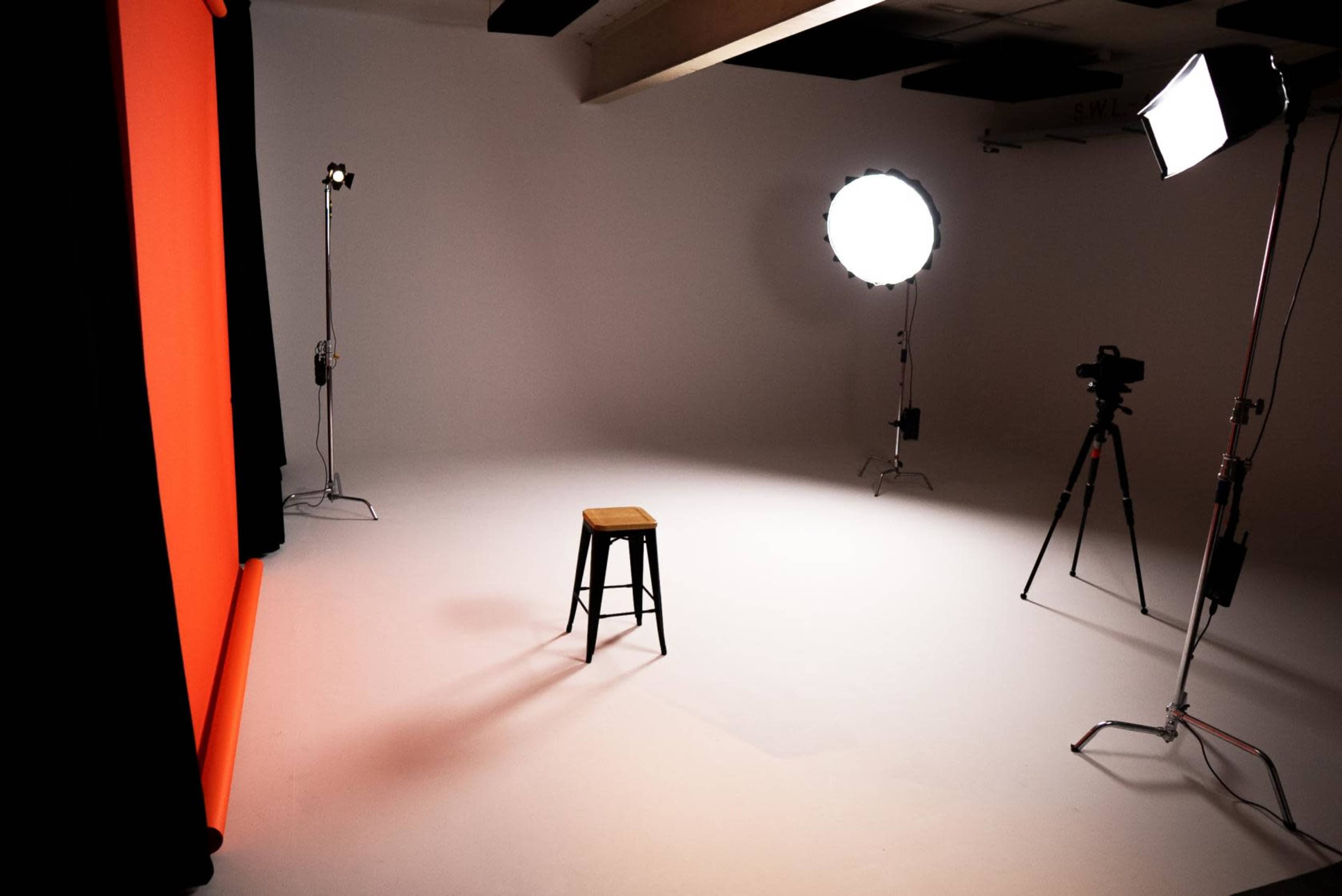 A photography studio with an empty wooden stool positioned against a plain background, flanked by various lighting equipment and a camera on a tripod.
