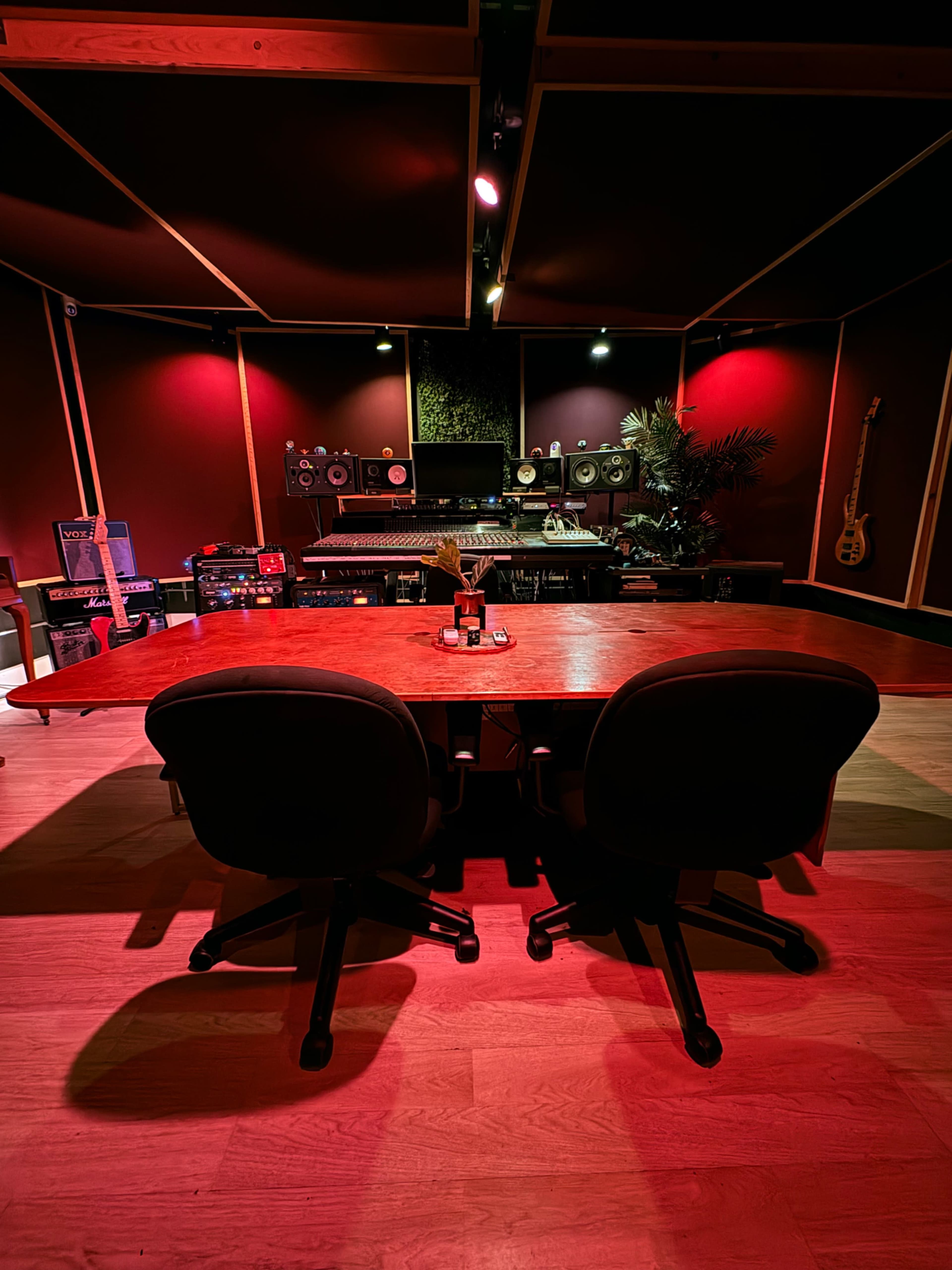 The image shows a music recording studio with two chairs at a wooden desk, surrounded by audio equipment and illuminated by red lighting.