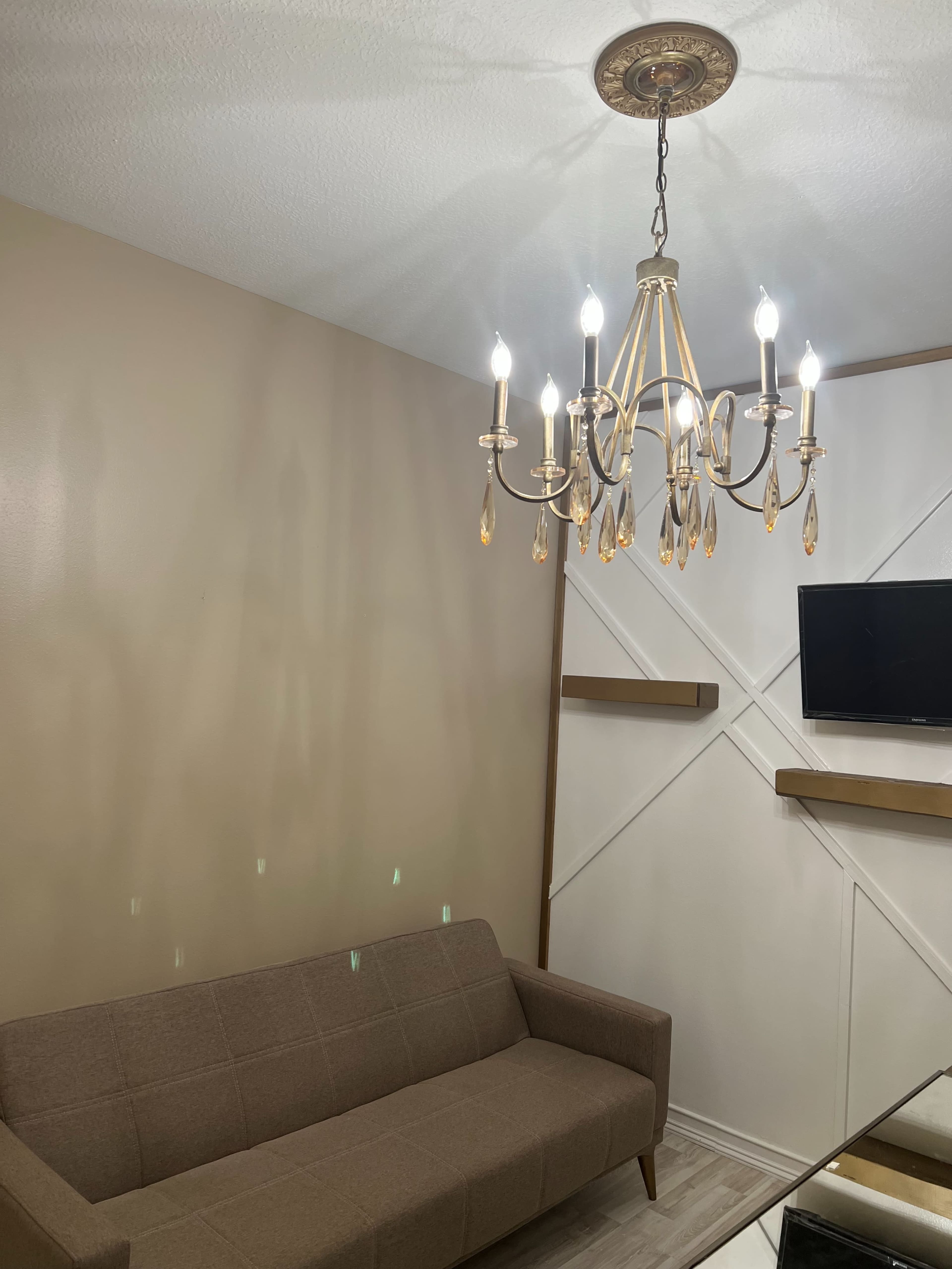 A chandelier hangs above a beige sofa in a room with light-colored walls and a mounted television.