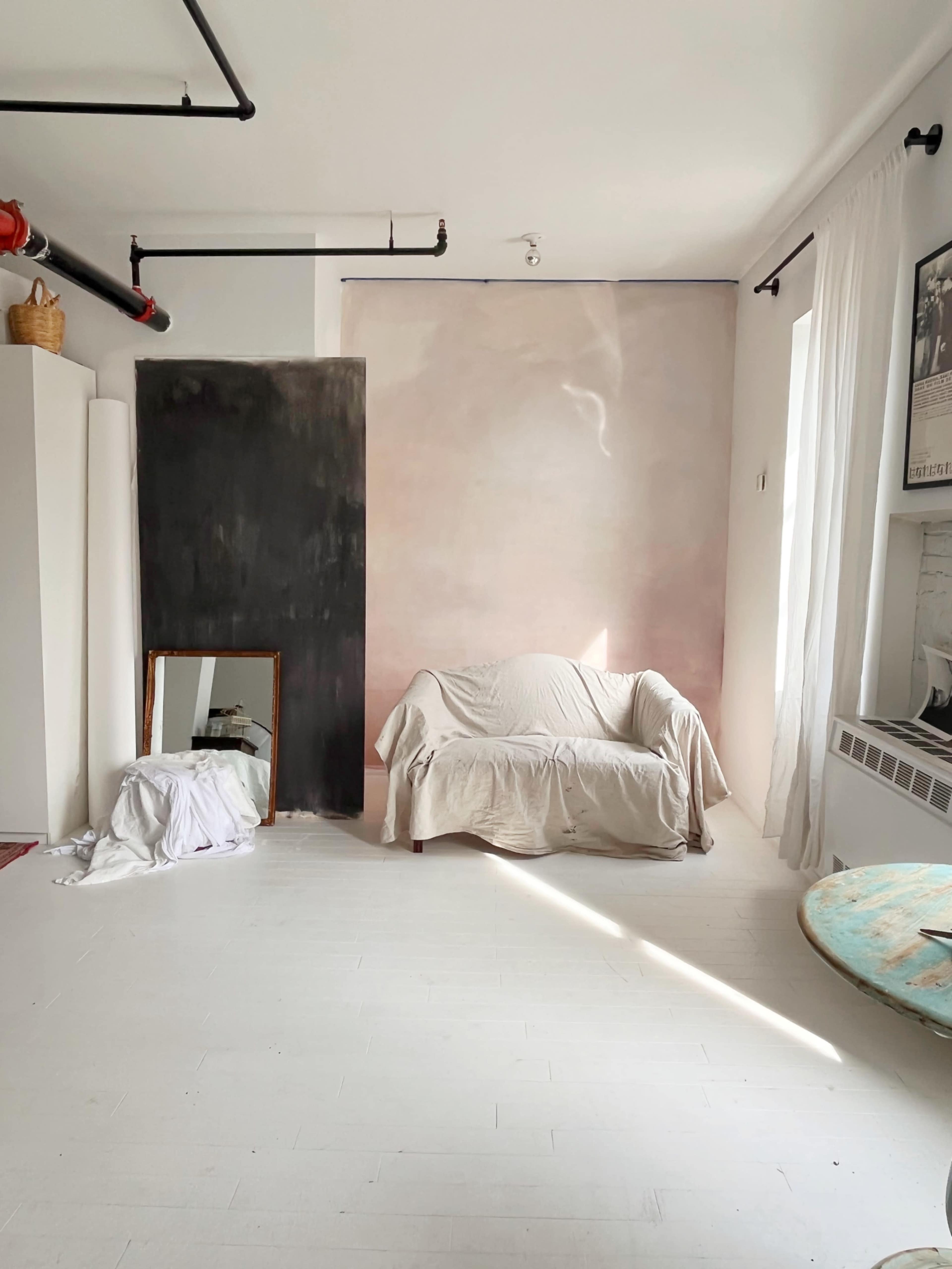 A minimalist room with a covered couch, a large mirror on the floor, and a wall painted in soft pink and dark gray tones, illuminated by natural light.