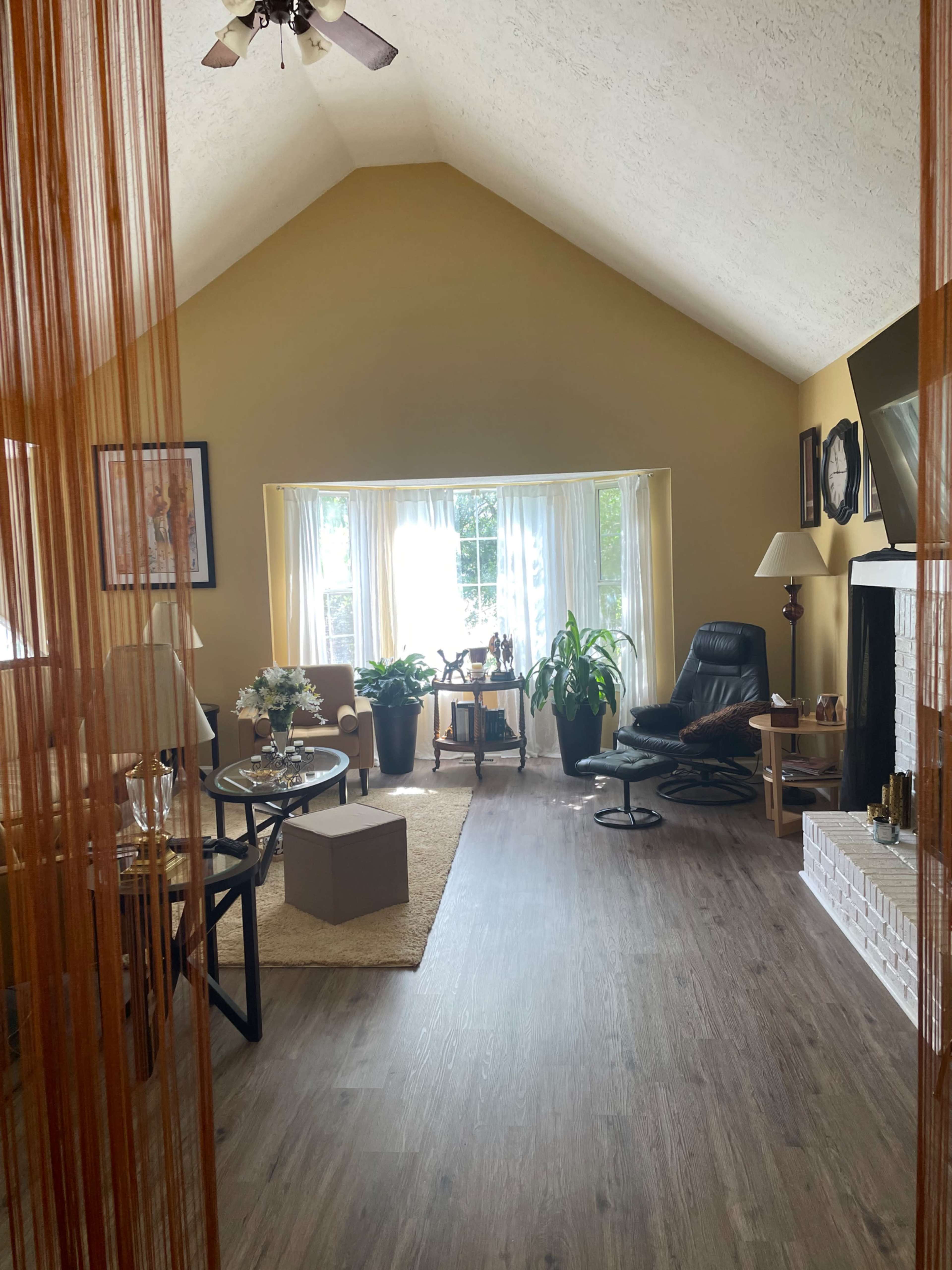 A spacious living room features a large window, plants, a black recliner, and light-colored walls with wooden flooring.
