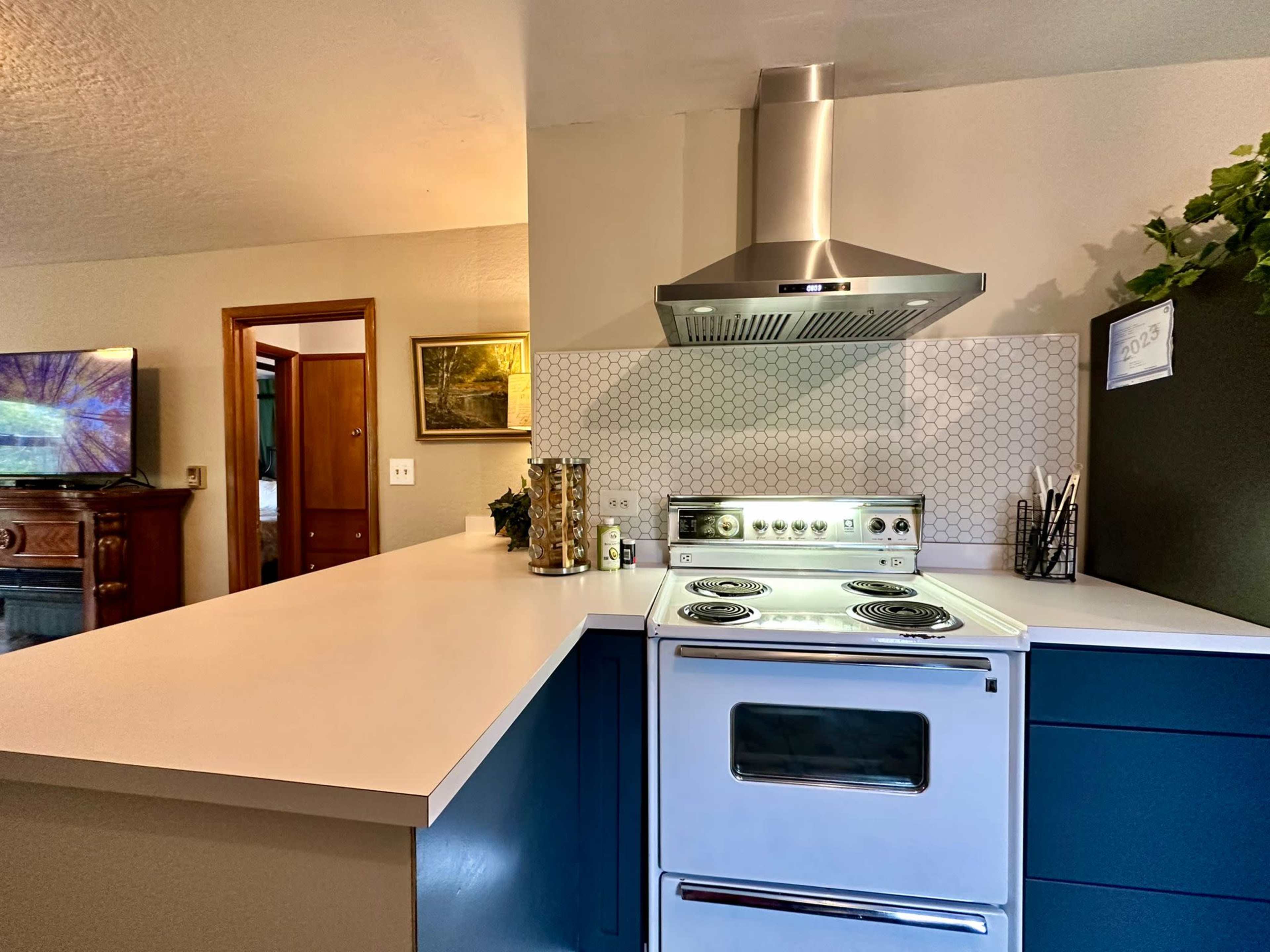 The image shows a modern kitchen featuring a white countertop, a blue cabinet, a stainless steel range hood, and a vintage-style white stove.