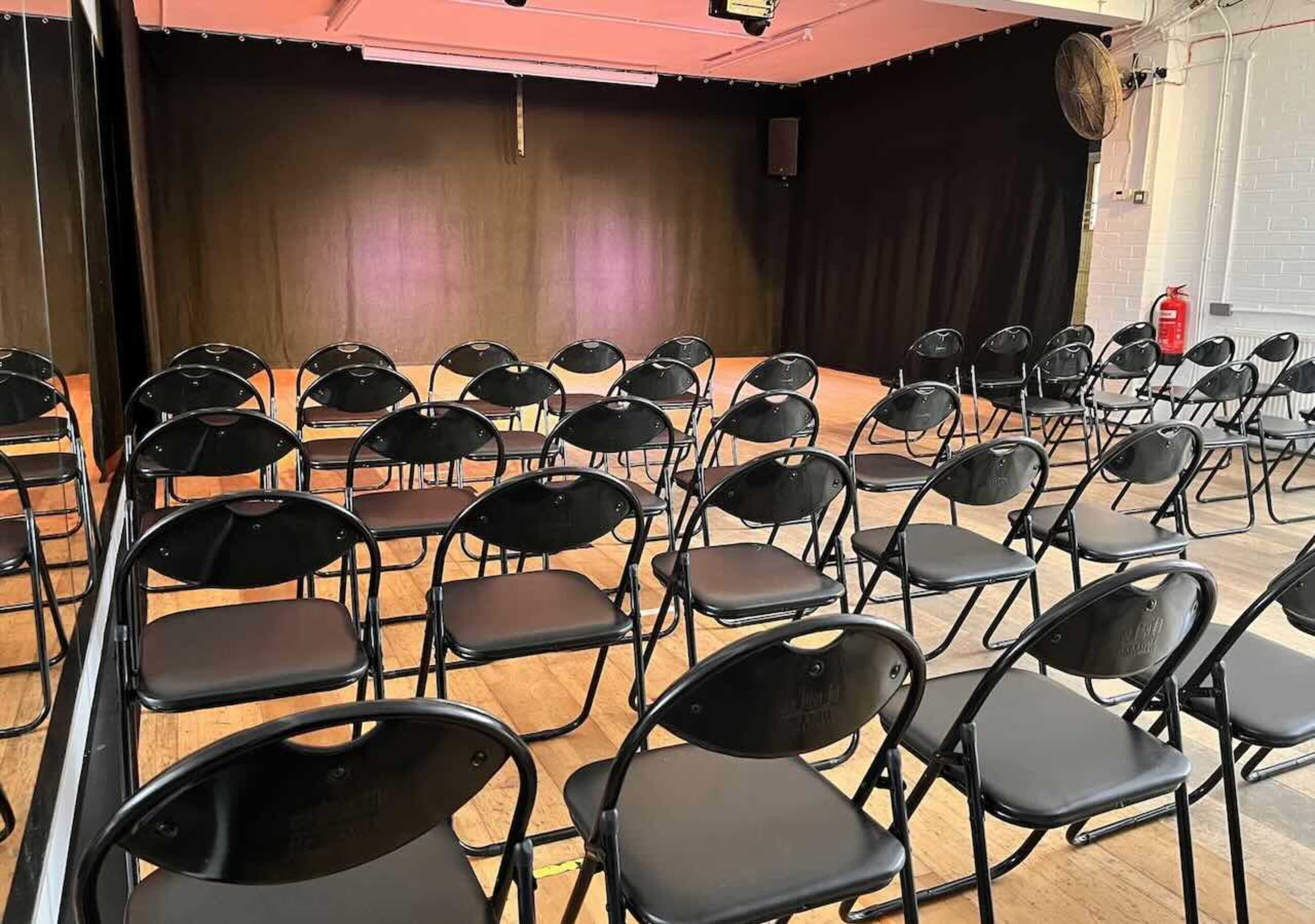 The image shows a venue with a stage at the front and rows of black folding chairs arranged in front of it.